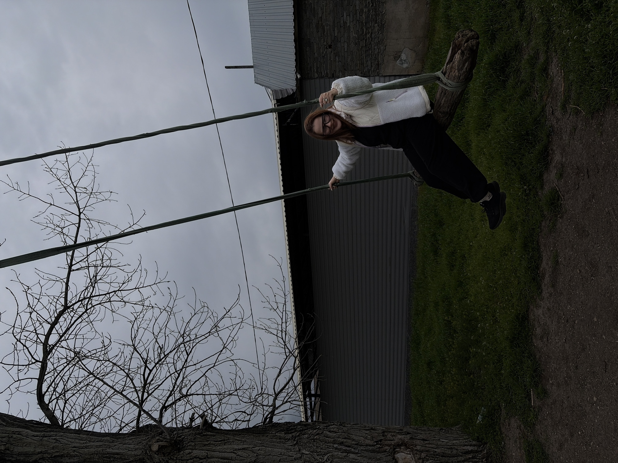 A young woman with glasses is on a swing with a wooden seat, in an outdoor location. She wears a white jacket and dark pants, and black shoes. The swing is suspended from ropes attached to a tree branch. Behind her is a building with a gray roof and gray wall, and a patch of green grass. The sky is overcast. The location is likely a recreational area in Port-Katon, Russia, considering the user prompt.