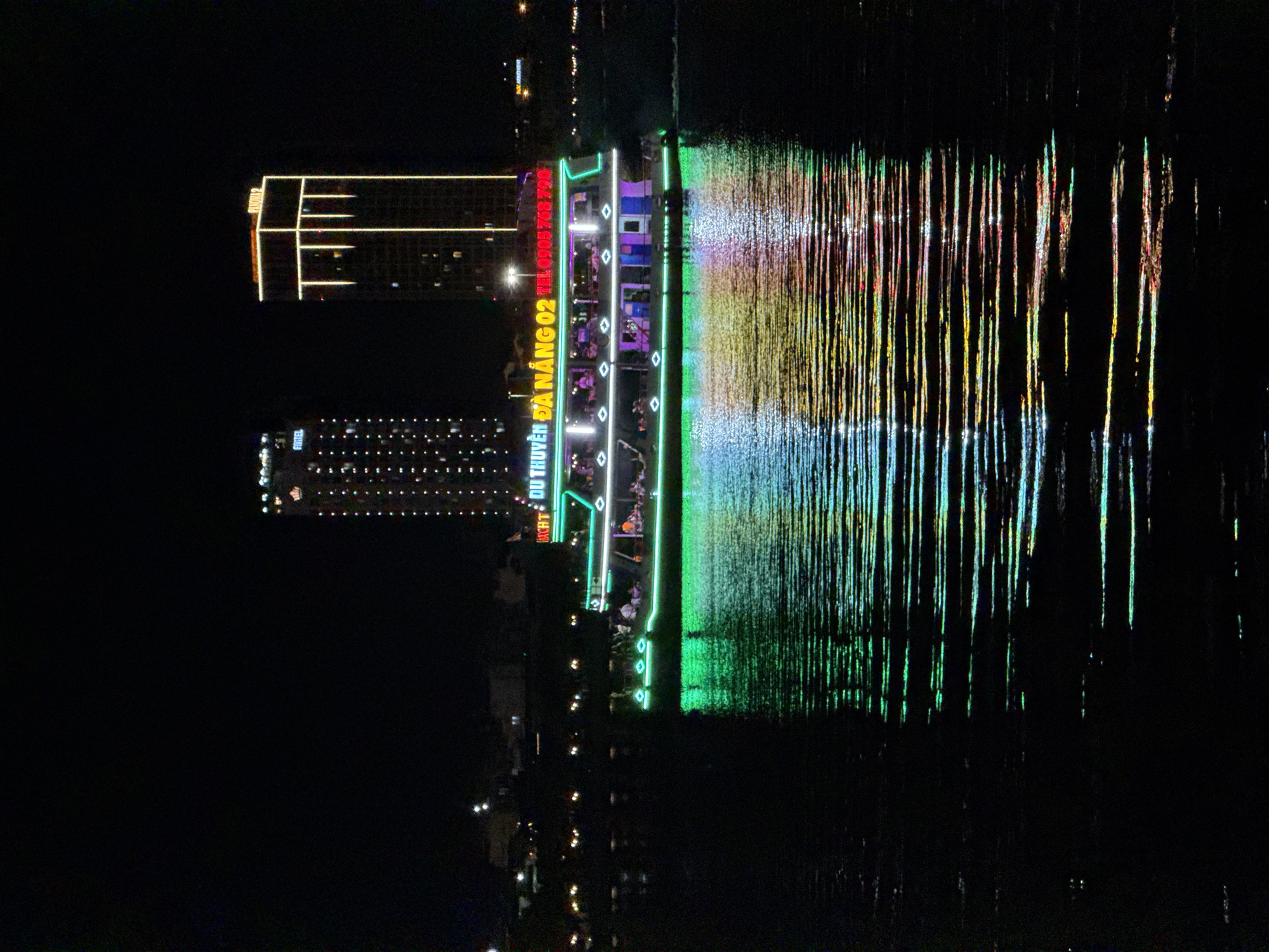 The image captures a nighttime scene on the Hàn River in Đà Nẵng, Vietnam. A two-deck passenger cruise boat, labeled "DU THUYỀN ĐÀ NẴNG 02" (DA NANG CRUISE 02) with a contact number, occupies the central frame. The boat is brightly illuminated with green LED strips along its lower hull and perimeter, white lights within its passenger cabins on both decks, and pink/purple accent lighting on the lower deck sides. Multiple passengers are visible on both decks, seated at tables. The boat is moving, indicated by a faint wake behind it.
In the background, two illuminated high-rise buildings rise against a dark sky. The left building features numerous small, scattered lights and the faint text "HOTEL" at its summit. The right building is outlined with prominent yellow-gold light strips along its edges and vertical architectural features. Distant, indistinct city lights line the riverbank.
The boat's colorful illumination is reflected vividly on the rippled river surface, producing elongated, horizontally fragmented streaks of green, blue, yellow, orange, and red light directly beneath and extending to the right of the vessel. The water in the foreground is dark, save for these reflections.