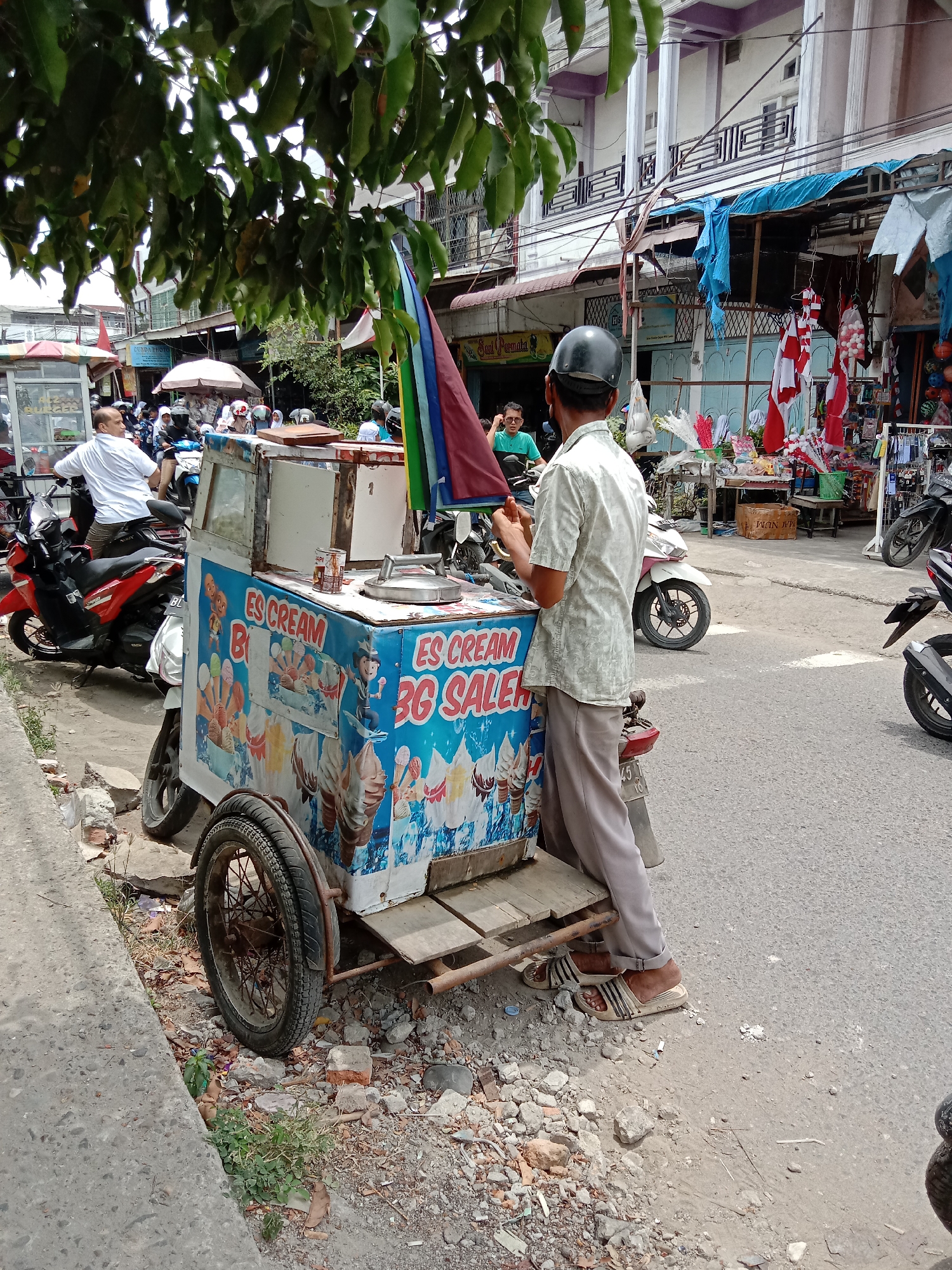 The image depicts a street scene in Lhokseumawe, Indonesia, with a focus on an ice cream cart. The cart is blue and decorated with images of ice cream cones, and text that reads "ES CREAM BG SALEH". A man wearing a light-colored short-sleeved shirt, gray pants, and a helmet is standing next to the cart, appearing to be interacting with it or its contents. He is wearing sandals. Several motorcycles are parked nearby, and other people can be seen in the background, suggesting a busy urban environment. The buildings in the background are typical of Indonesian street architecture, with multiple stories and balconies. The overall setting is a street with a paved road and a concrete sidewalk. Trees with green leaves provide shade over part of the scene. The lighting suggests it is daytime.