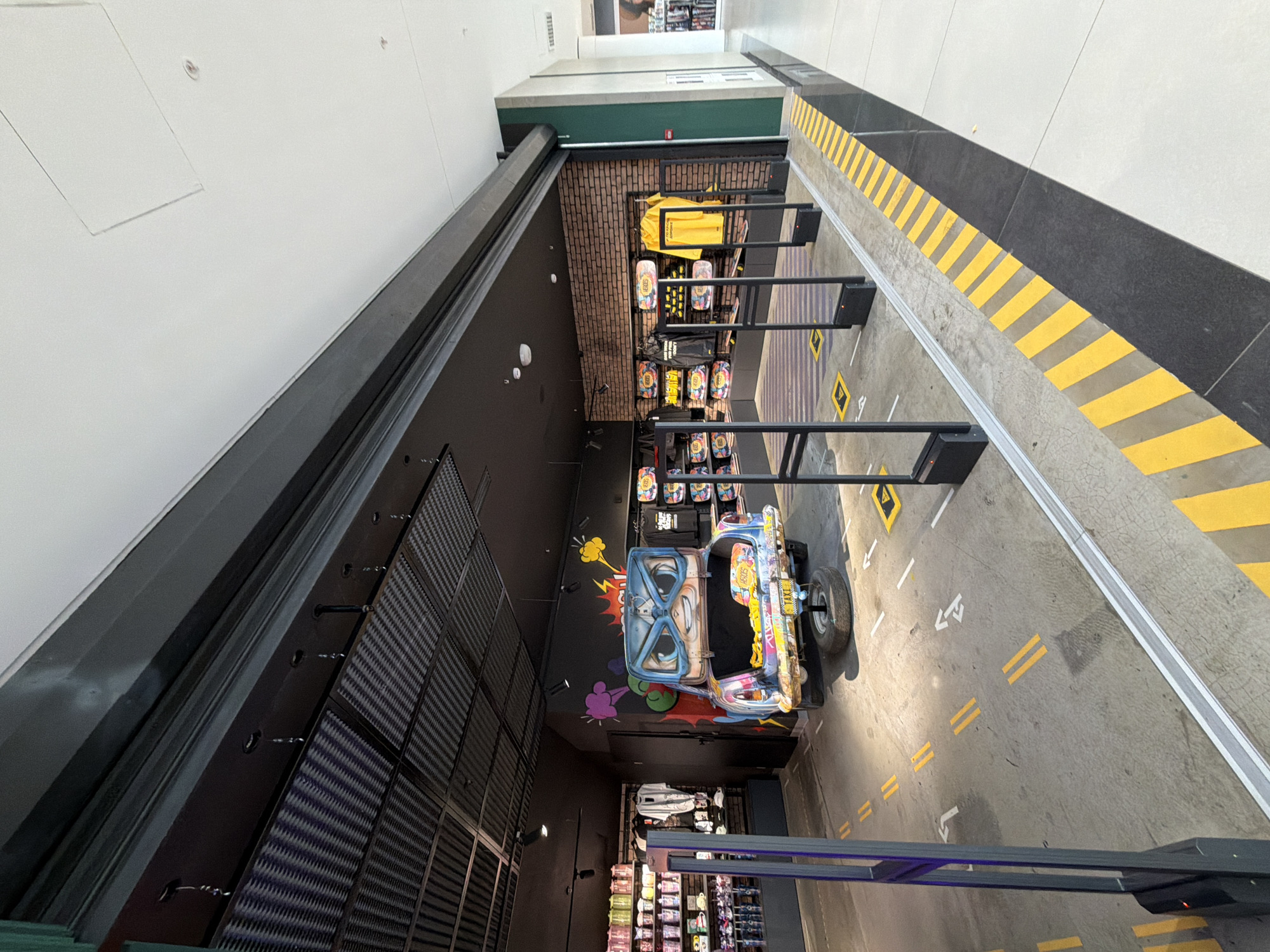 This high-angle image depicts an indoor commercial space, likely a retail store or a themed entertainment area, possibly located in Minsk, Belarus. The setting features a playful, urban street-style design, viewed from above.
In the foreground, a vibrant, customized mini-vehicle, resembling a buggy or jeep, is parked on a grey concrete-like floor. The vehicle boasts a large, cartoon-style face painted on its front, complete with blue-rimmed glasses, and its body is covered in colorful graffiti-like graphics. "TAXI" is visible on a rear license plate.
The grey floor beneath the vehicle and extending into the midground is marked with yellow and white road lines, including dashed center lines, double yellow lines, and directional arrows, simulating a street. A yellow diamond-shaped warning sign with an exclamation mark is also painted on the floor.
To the right, a wide strip of floor features prominent yellow and black diagonal stripes, indicating a hazard or border zone, adjacent to a darker, possibly black, flooring section. To the left, a dark wall is covered with textured, square panels, while overhead, lighter walls and a ceiling suggest a spacious interior, likely part of a larger complex like a shopping mall.
Various retail displays are scattered throughout the area, showcasing brightly colored merchandise such as bags, skateboards with graphic designs, and a vivid yellow garment. Tall, dark metal barriers or security gates are also part of the setup. In the background, a brick-patterned wall adds to the urban aesthetic, situated beneath a light-colored structure with a dark green base. The scene is well-lit, characteristic of an indoor commercial environment, and no people are visible, presenting a static display of the space.