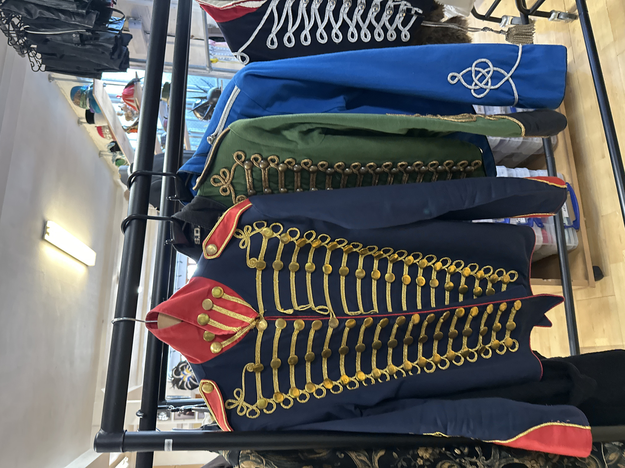 The image depicts a rack of military-style jackets on display, likely within a store or museum. The jackets vary in color, including navy, blue, and green, and are heavily adorned with gold embellishments and button detailing. The setting appears to be indoors.