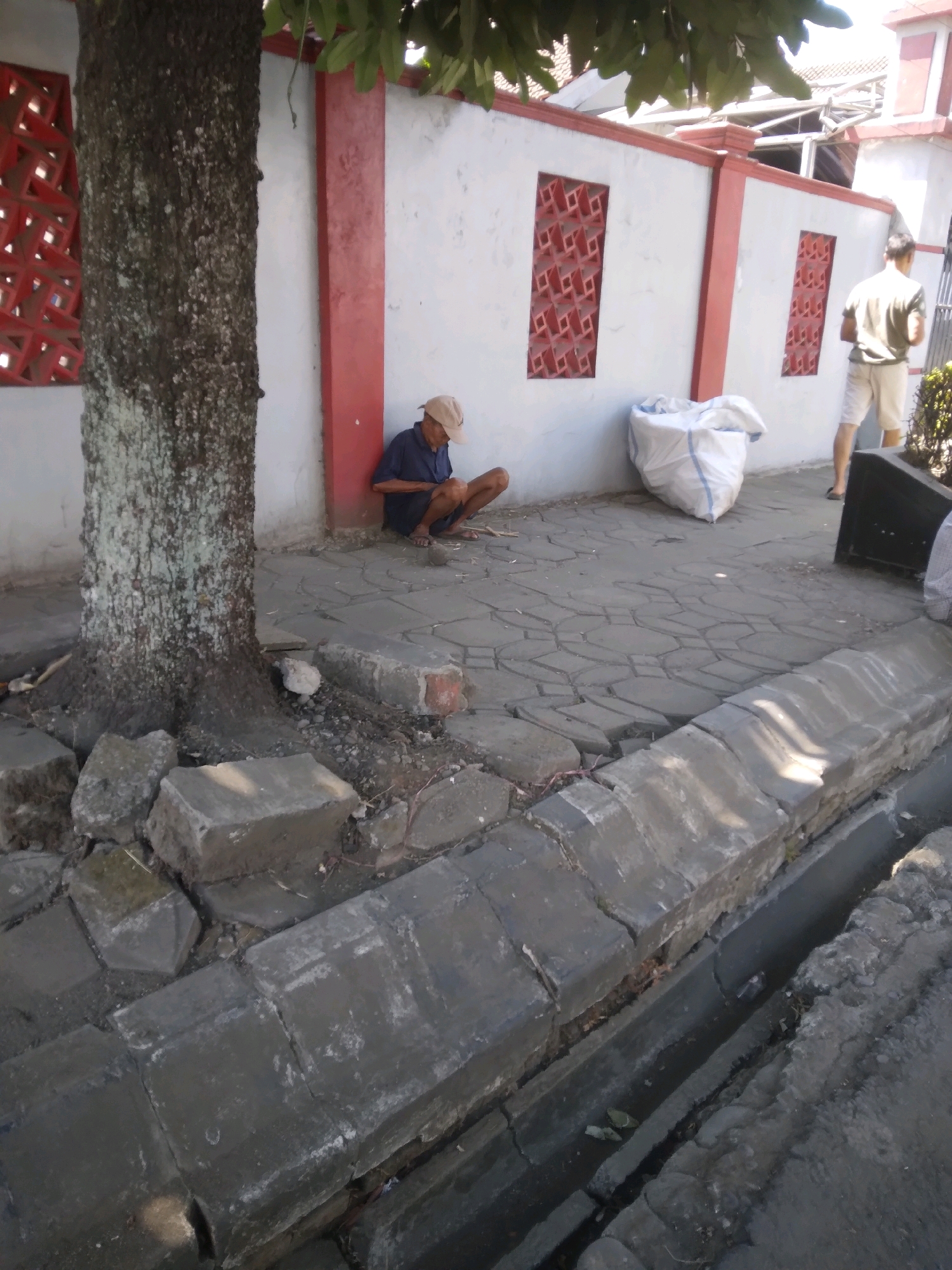 The image depicts a sunlit outdoor scene, likely a street or walkway in Sumampir, Indonesia, characterized by cracked pavement and a bordering drainage ditch. A large tree trunk occupies the left side of the frame. In the middle ground, a man in a blue shirt and beige cap squats on the ground, looking down. Beside him is a white, large plastic bag. To the right, another man stands wearing a light brown shirt and white shorts. The background features a white wall with red decorative elements and pillars. Debris, including broken concrete blocks and stones, is scattered on the ground, particularly around the base of the tree and along the ditch.