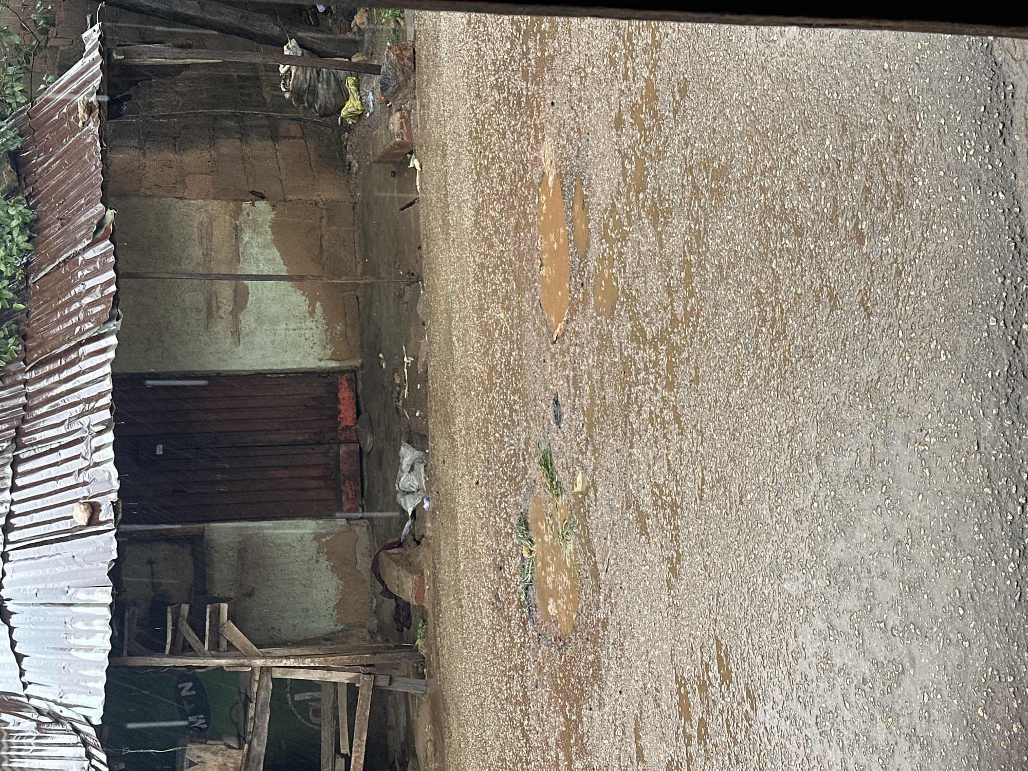 An outdoor, eye-level shot captures a weathered corrugated metal roofed structure with rough-hewn wooden supports, its walls constructed from mud-brick and cement with patches of white and brown. To the left of the entrance, a wooden frame supports what appears to be a partially obscured sign or advertisement. The ground in front of the structure is a dirt road, wet and pocked with several puddles reflecting the overcast sky. Scattered debris, including plastic bags and what looks like a sack of produce, is visible near the building's base. The scene is set in an urban environment, possibly a developing area, given the construction materials and the overall state of the road and building.