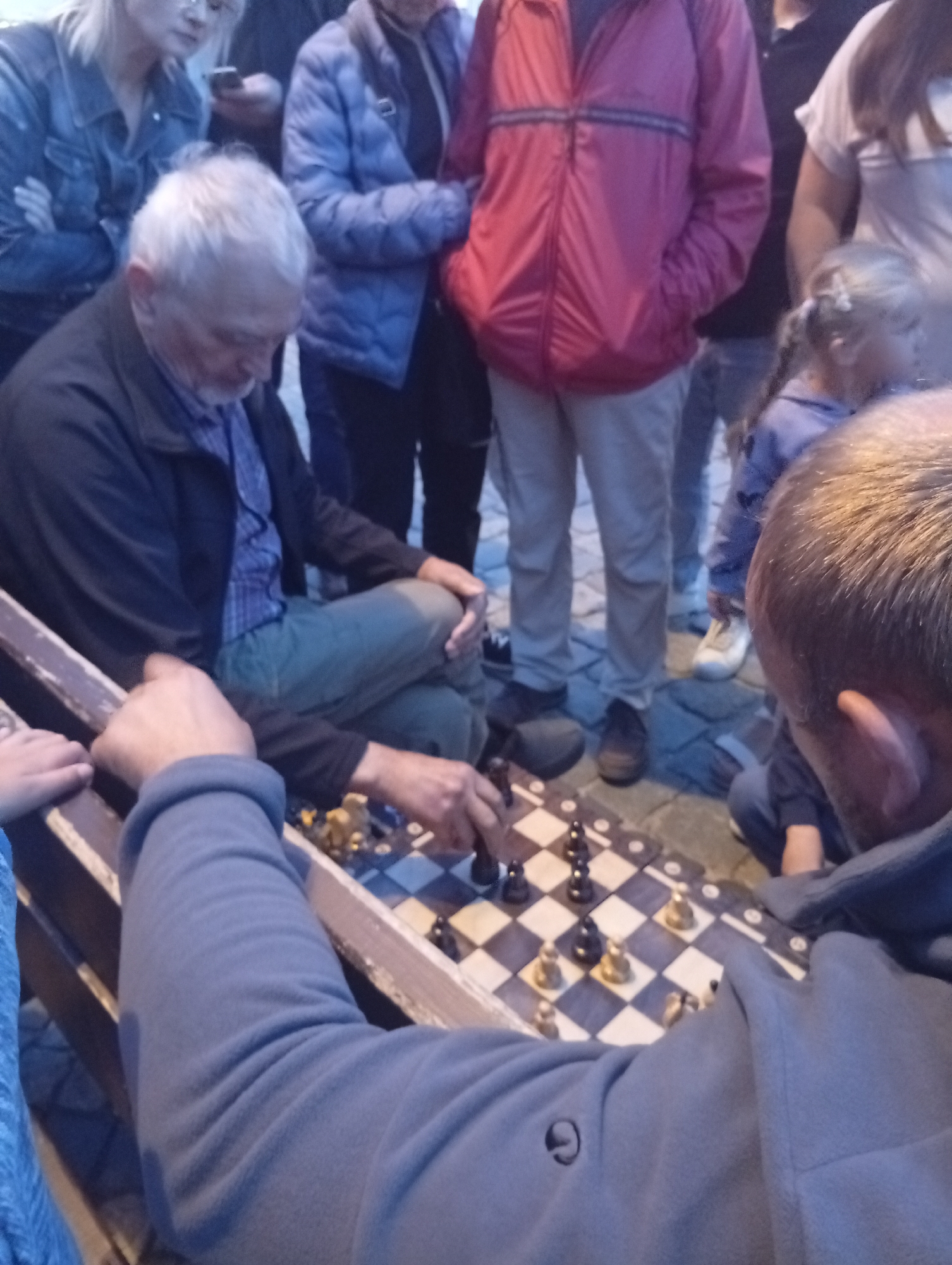 In Wrocław, Poland, a group of people are gathered around a chessboard laid out on a stone-paved area. An older man with gray hair and a blue checkered shirt is seated on a wooden bench, focused on the game. His legs are crossed, and he wears a dark jacket and green pants. In front of him, a chessboard displays a game in progress, with black and gold pieces. Two other individuals are partially visible, one on the right with their head and shoulder in the foreground, wearing a gray hoodie, and another person on the left. Several other people stand in the background, some wearing jackets in various colors, including blue and pink, and a young girl with blonde hair is also present. The scene appears to be outdoors, possibly in a public square or park.