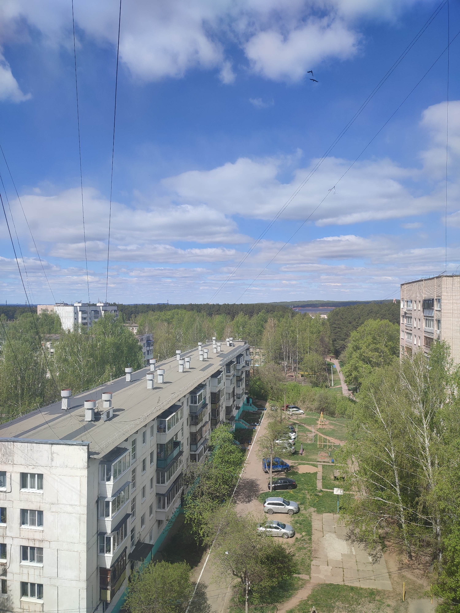 The image depicts an elevated view of an urban landscape, likely from an apartment building. The scene includes a residential area with apartment blocks, a park, and a glimpse of a body of water in the distance. The sky is blue with scattered clouds, and a couple of birds are visible. Electrical wires are strung across the foreground of the sky. In the middle ground, a row of apartment buildings is prominent, with a concrete roof and rows of balconies. Adjacent to the buildings is a park or green space with trees, a playground, and a parking area with several cars. The overall atmosphere suggests a quiet, residential area with access to green spaces and a view of the surrounding landscape.