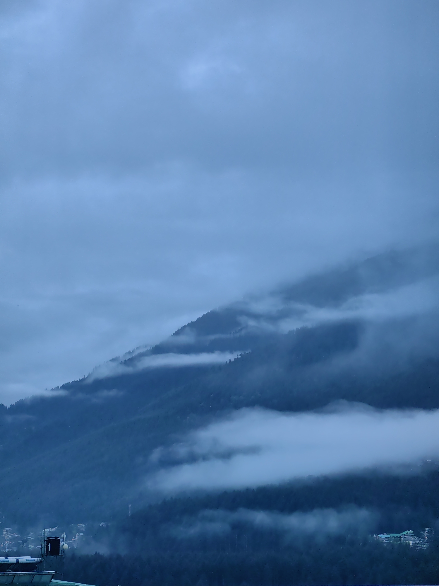 The image depicts a panoramic view of a mountainous landscape shrouded in heavy, grey clouds. Dense, dark green coniferous trees cover the slopes of the mountains. Wisps of white fog and low-lying clouds drift through the valleys and along the mountain ridges, obscuring parts of the terrain and creating a mystical, ethereal atmosphere.
In the lower part of the image, a section of the town of Vashisht, India, is visible. Buildings with sloped, green roofs are clustered together on the mountainside. Some of these structures appear to be hotels or guesthouses, typical of a mountain resort town. In the foreground, parts of a building's roof are visible, along with a black water tank, and what appears to be a communication tower. The overall scene suggests a cool, possibly damp, day with limited visibility due to the weather conditions.