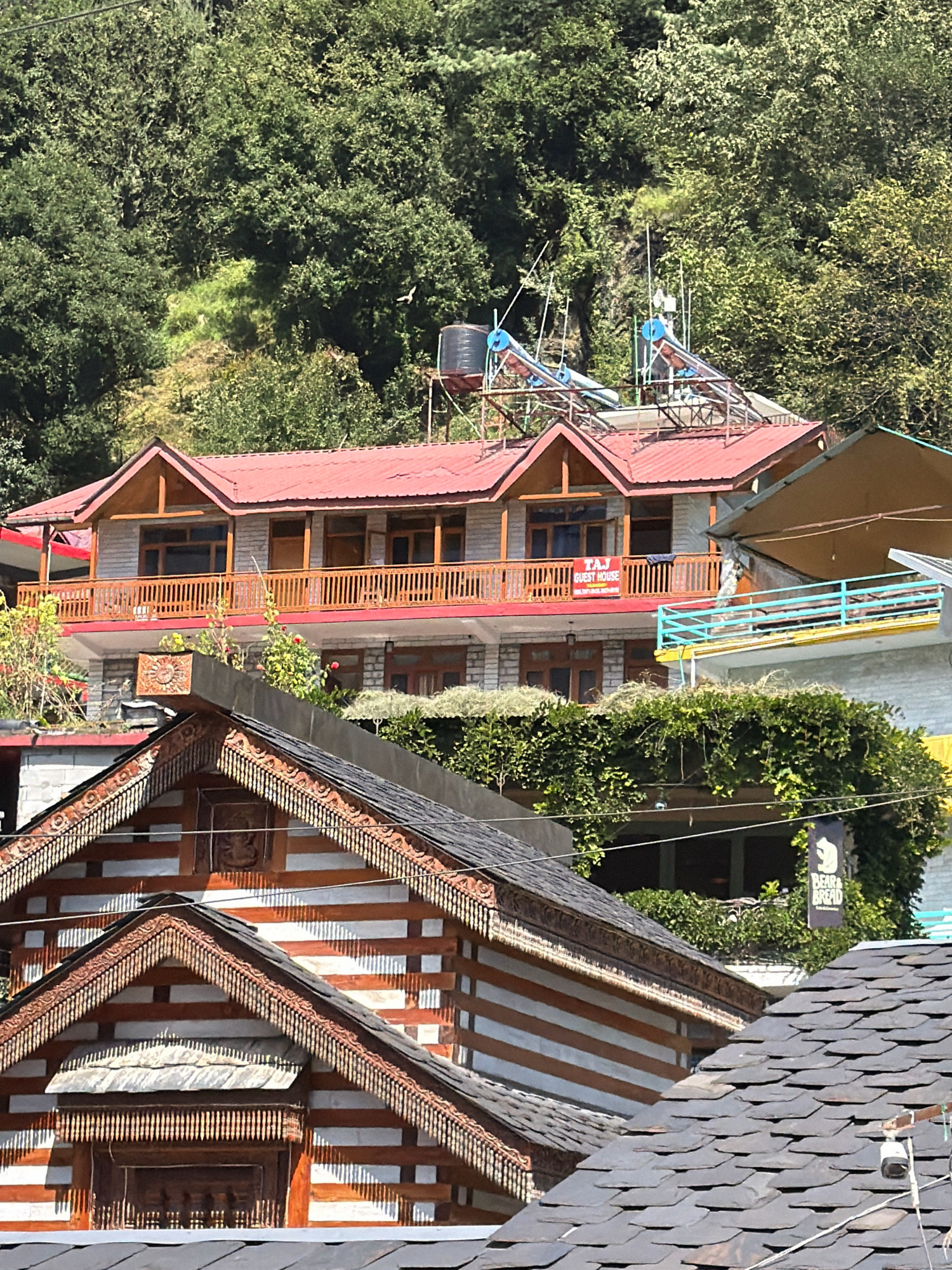 The image is a photograph of buildings in Vashisht, India, taken on a sunny day. In the foreground, there are several roofs with dark grey slate tiles and traditional wooden architectural elements. Further back, a multi-story building with a red metal roof is visible. This building features wooden balconies with railings and stone walls. A sign reading "TAJ GUEST HOUSE" is attached to one of the balconies. On the roof of this building, there are two solar water heaters. Behind the buildings, a dense, green forest covers a hillside.