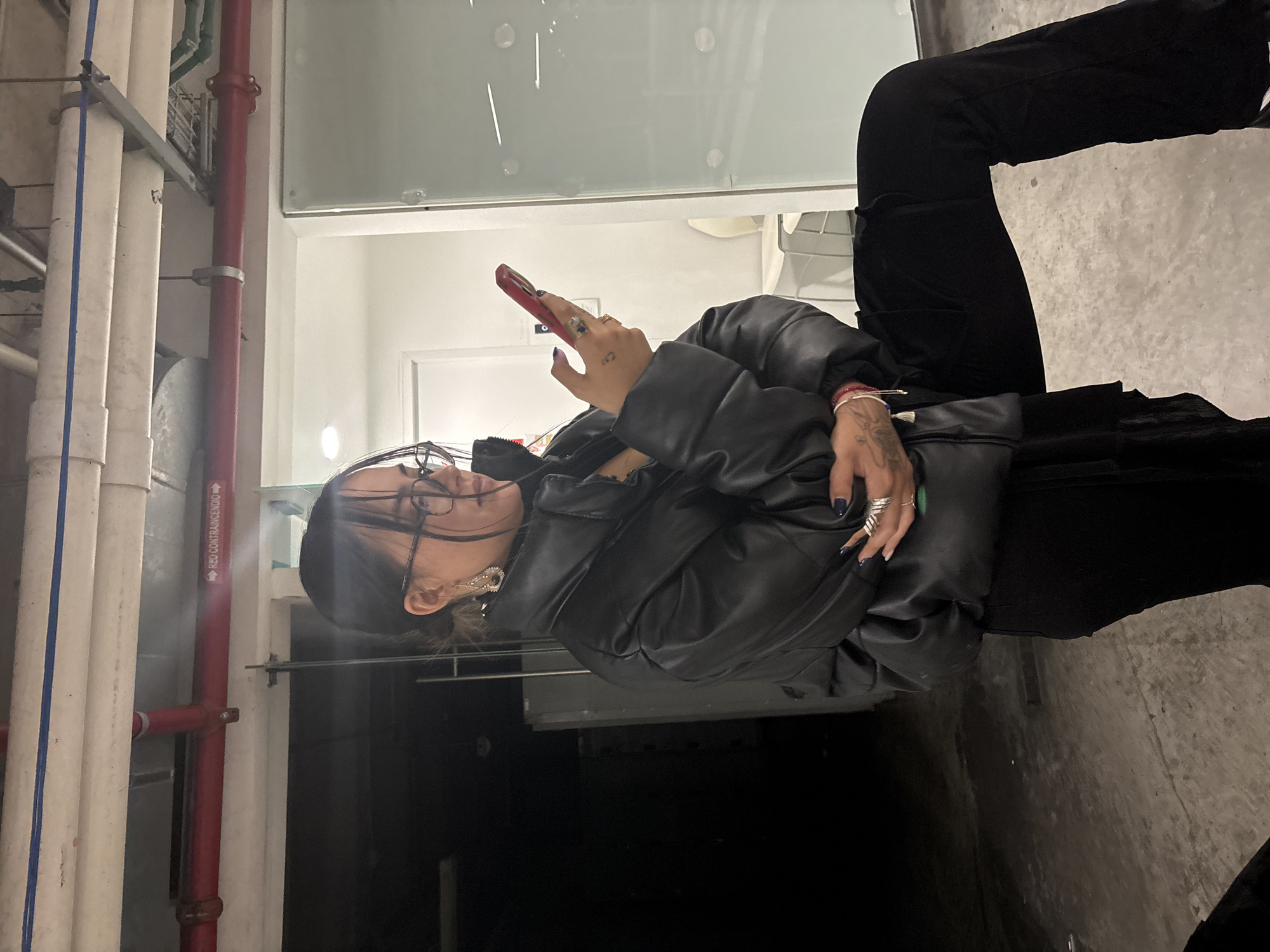 A person, likely a woman with dark hair and glasses, is captured in a vertical, seemingly candid shot, intently looking down at a red smartphone held in her hands. She is dressed in a black, possibly puffer-style, leather jacket and black pants. Several rings adorn her fingers, and a tattoo, resembling a small heart or loop, is visible on her right hand. Distinctive earrings hang from her ears.
The setting appears to be an indoor or semi-enclosed utility space, characterized by exposed industrial piping on the left side and top of the frame. White pipes run vertically, alongside a prominent red pipe. On this red pipe, the Spanish text "RED CONTRAINCENDIO" (meaning "FIRE NETWORK" or "FIRE FIGHTING NETWORK") is clearly visible, providing a strong contextual clue for a Spanish-speaking region, consistent with Mexico City. The background also includes a light-colored wall with a white door and what appears to be a glass partition or window, suggesting an adjacent, possibly brighter area. The floor is a textured concrete surface. The person is focused on her phone, seemingly engrossed in an activity, and her expression is neutral and concentrated.