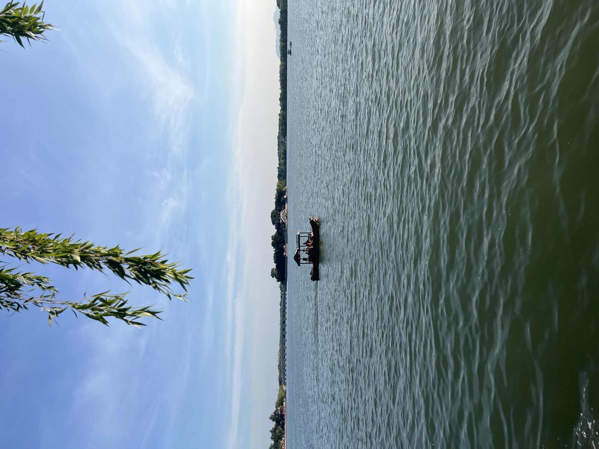 The image shows a wide view of a lake on a clear day with wispy clouds in the sky. In the foreground, green leaves hang from a tree branch, partially obscuring the upper portion of the frame. In the middle ground, a small boat with a canopy is on the water, carrying several passengers. In the background, the shore is lined with trees and some traditional architecture, including a long, arched bridge. Another small boat is visible in the distance on the right side of the lake. The water is calm with gentle ripples.
