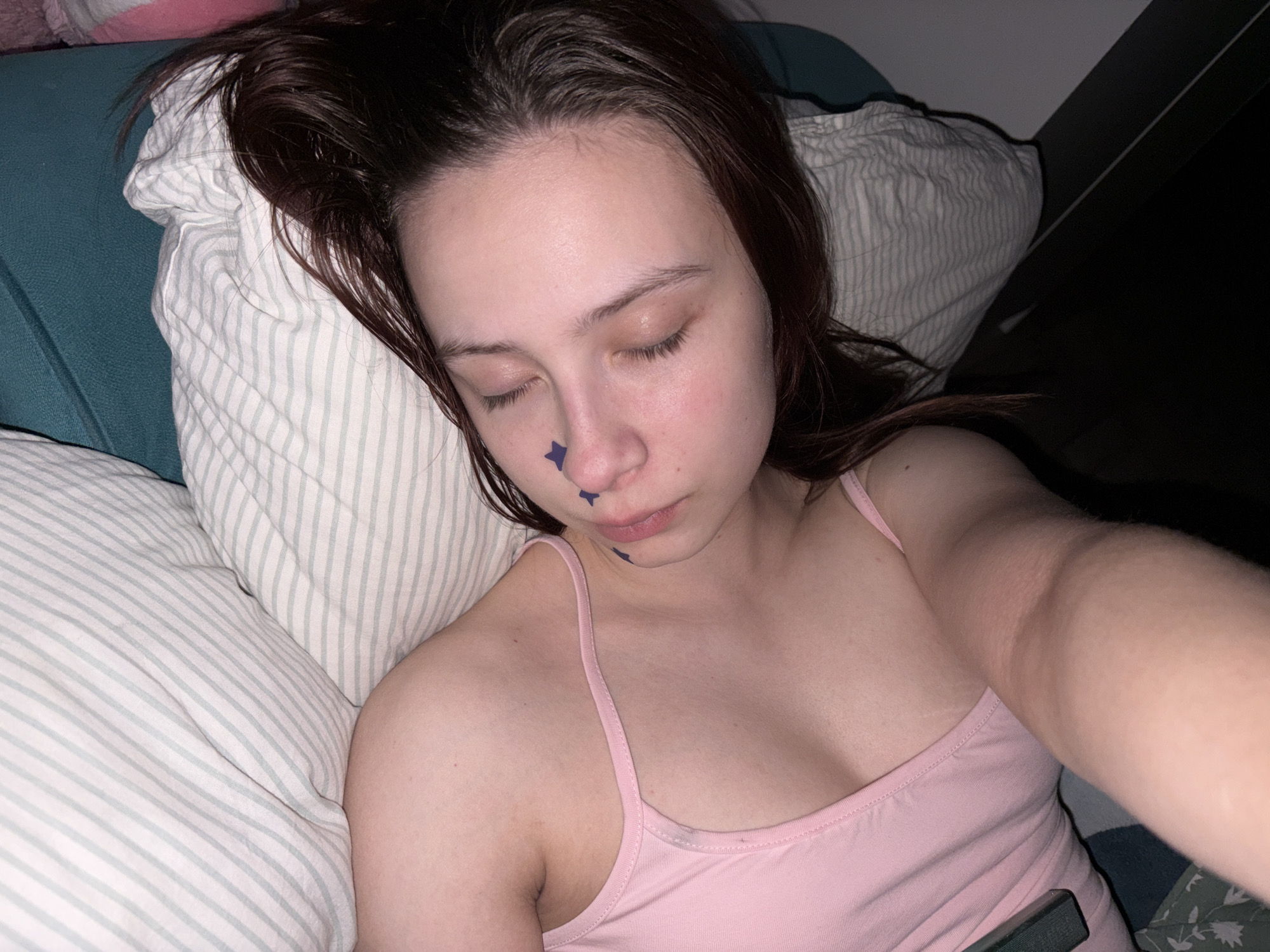 A young woman with light skin and brown hair is lying on a bed. She is wearing a pink tank top. There are two blue star-shaped stickers on her face, one on her nose and the other on her upper lip. Her eyes are closed. She is lying on a pillow with green and white striped fabric. There is a dark object, possibly a phone, near her right arm. The background is dark with a glimpse of a teal-colored object. There is no indication of the city of Прикамский, Russia, in the image.