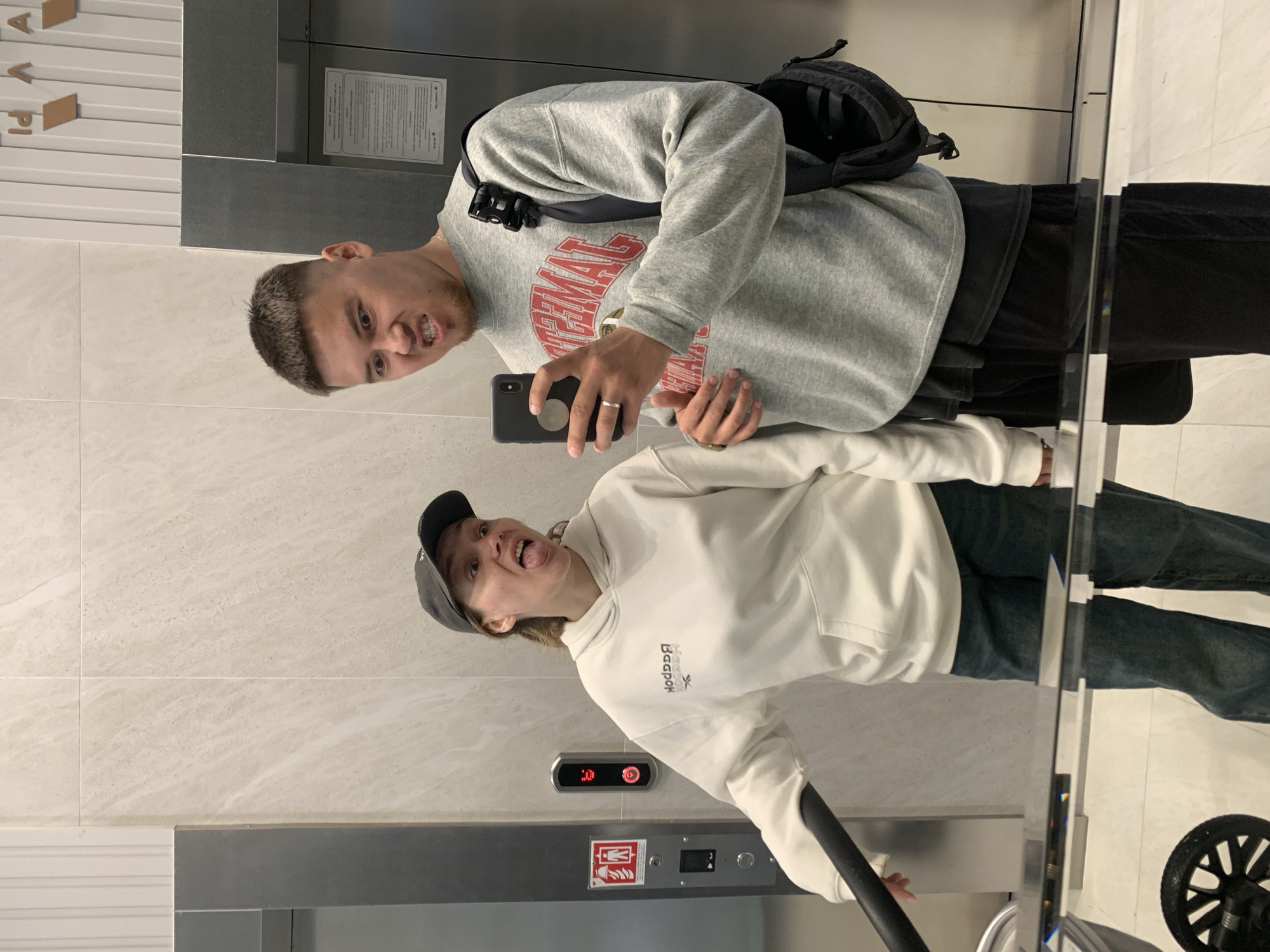 In an elevator in Stavropol, Russia, a young man and woman are taking a mirror selfie. The man, wearing a grey sweatshirt with red lettering and a black crossbody bag, is making a grimace at the camera. The woman, in a white hoodie and a black baseball cap, sticks her tongue out playfully. The elevator walls are tiled with a light marble pattern, and a silver handrail is visible in the foreground. A stroller is partially visible to the left. The floor is tiled with a light grey pattern, and a reflective surface, likely the elevator doors or a mirrored wall, captures their reflection. An elevator call button panel with a red light is mounted on the wall.