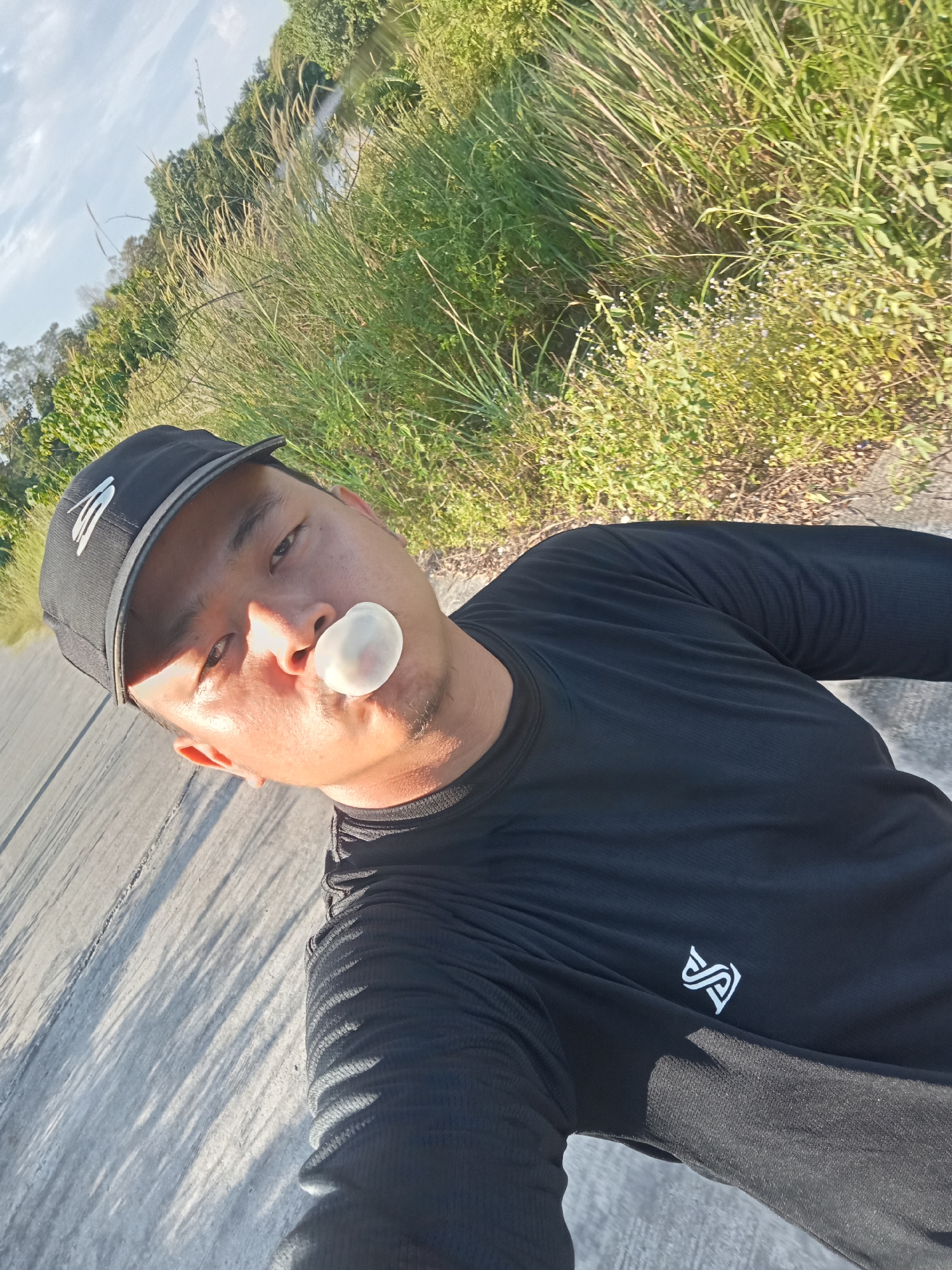 A close-up selfie shows a man wearing a black cap and a black long-sleeved shirt. He is outdoors, with a road in the foreground and lush green vegetation, including tall grasses and trees, bordering a body of water in the background. Sunlight casts long shadows across the road. The man has a bubble of chewing gum inflated in front of his mouth. The environment appears to be a rural or semi-rural area.