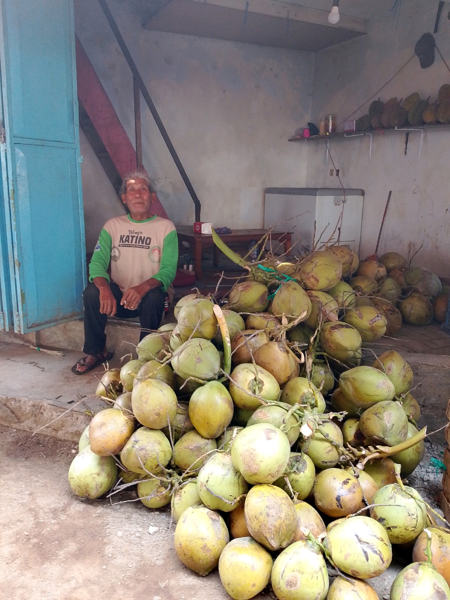A man sits on a step in front of a stall selling coconuts in Tiron, Indonesia. He is wearing a tan and green long-sleeved shirt with "KATINO" printed on it, and black pants. He is wearing sandals. In front of him is a large pile of green coconuts. Behind him, there is a blue door, a shelf with several durians on it, and a white refrigerator. The ground is dirt and gravel.