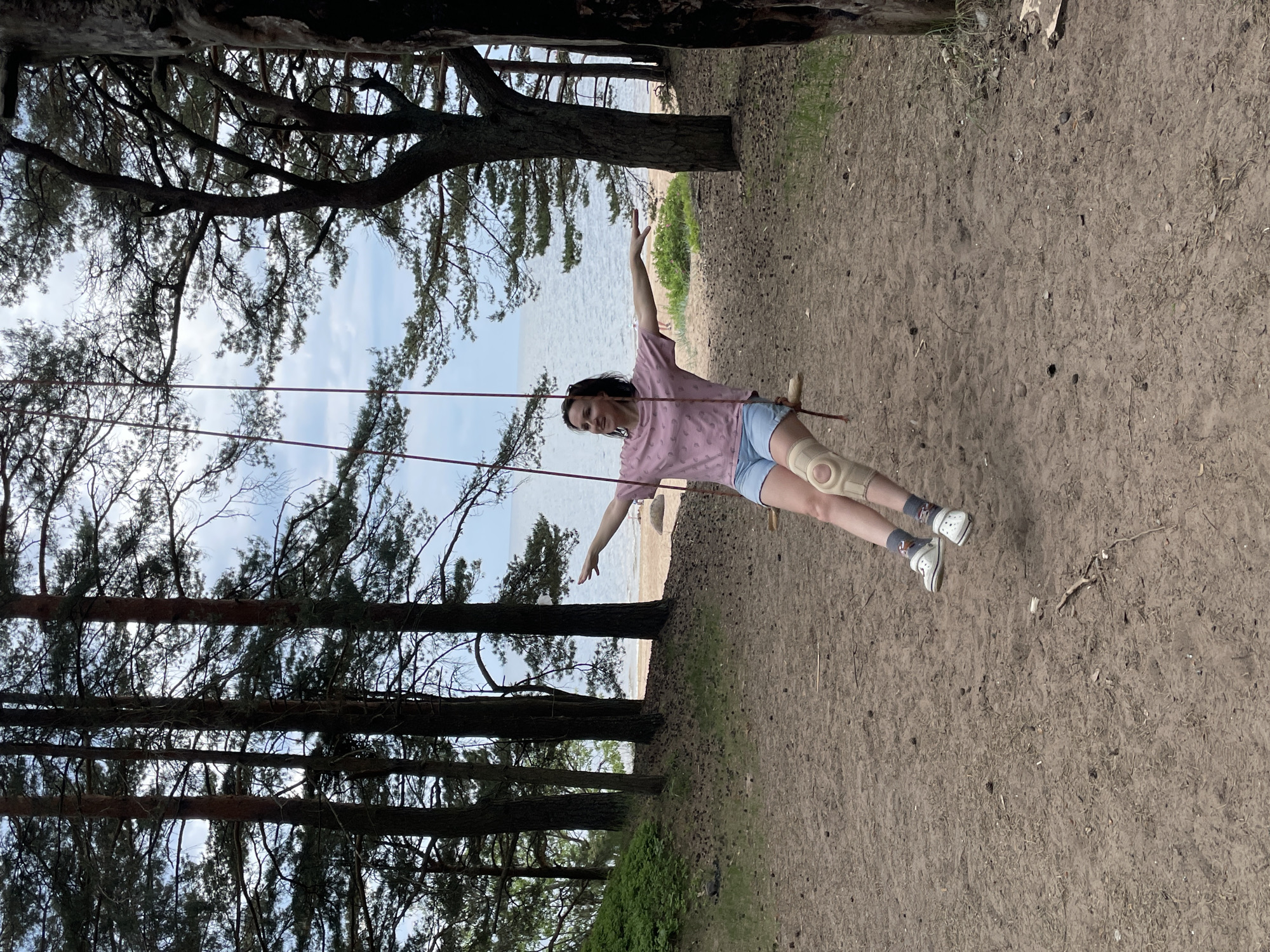 A woman is depicted mid-swing on a wooden swing with red ropes, positioned in a sandy, forested area in Zelenaya Roshcha, Russia. She is facing slightly to her right, smiling with her dark hair wet or slicked back, and her arms are extended horizontally outwards. She wears a light pink short-sleeved t-shirt with a subtle pattern, light blue denim shorts, dark patterned socks, and white clogs. A beige knee brace with a patella opening is visible on her left knee. The ground is covered in sand with scattered pinecones and sparse vegetation. Behind the woman, a dense grove of tall pine trees with dark trunks and green needles separates the immediate foreground from a sandy beach and a calm body of water extending to the horizon under a partly cloudy sky. To the far right, the trunk of the tree supporting the swing is partially visible.