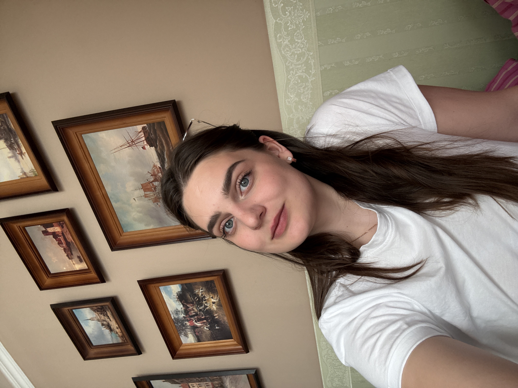 A portrait-oriented photo features a young woman looking directly at the camera, taken in what appears to be an indoor setting in Trnava, Slovakia. She is positioned diagonally across the frame, with her head and upper torso visible, and her long, dark brown hair flowing down her back and over her shoulders. Her blue eyes meet the viewer, and she has a gentle, calm smile on her face. She wears a plain white short-sleeved t-shirt, and a thin, light-colored necklace is visible around her neck. A small, sparkly earring is in her right ear, and a pair of eyeglasses rests on top of her head.
The background consists of a light beige or tan wall adorned with several framed artworks. These paintings, in various sizes and dark wooden frames, depict traditional scenes, including sailing ships at sea and what appear to be cityscapes or landscapes. A patterned light green and white wallpaper or fabric is visible to the right behind her, suggesting a sofa or upholstered furniture. A striped pillow, in shades of pink and white, is also partially visible on the far right. The lighting suggests it is daytime, with soft, even illumination. No discernible text is present in the image. The overall atmosphere is relaxed and homely.