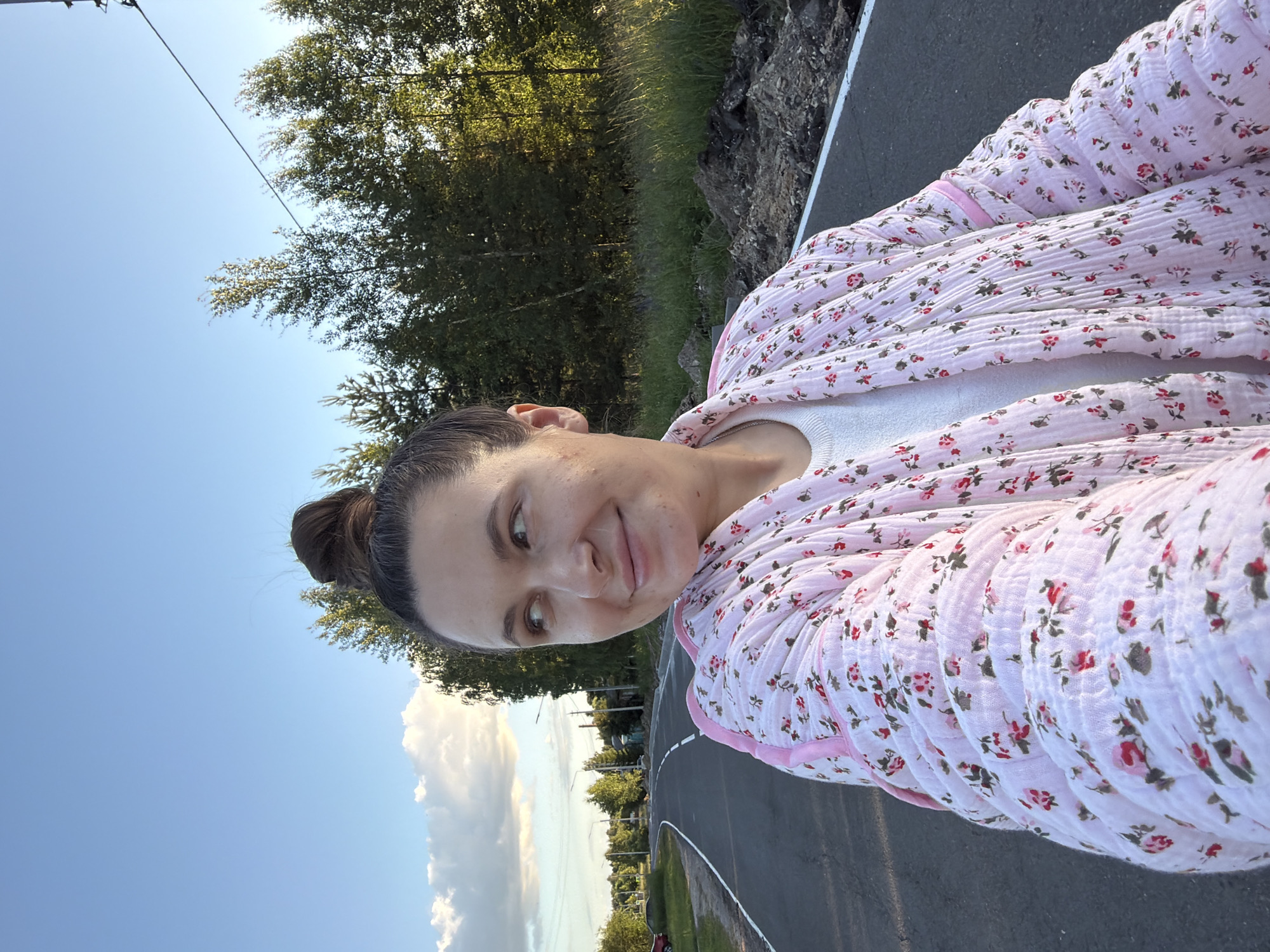 A woman is captured in a close-up, selfie-style shot. She has her hair pulled back in a bun and is smiling at the camera. She is wearing a light-colored, floral-patterned robe over a white top. The background shows a clear blue sky with some white clouds, trees, and a road. The lighting suggests it is daytime, likely late afternoon or early evening due to the golden hue of the light. The setting appears to be outdoors, possibly near a park or a rural road, given the presence of trees and grassy areas. The woman's expression conveys happiness and a relaxed mood. The image is oriented vertically, as if taken with a phone held in portrait mode. The exact location is specified as Красный Маяк, Russia.