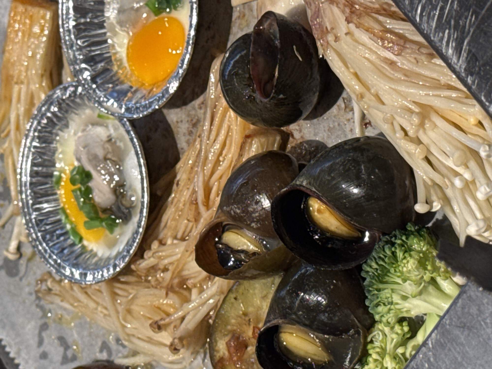 The image presents a close-up of food items arranged on a surface, possibly in a restaurant or a market setting. Two small, round, foil-like containers hold what appear to be egg yolks, one with additional green garnish and what could be an oyster. To the side, a cluster of dark-shelled snails occupies the central space. Enoki mushrooms appear in two different forms: one is a yellowish, cooked mass, and the other is fresh, white, and separated. A small piece of green broccoli is also visible. The scene is suggestive of Vietnamese cuisine, and considering the prevalence of seafood dishes in the city, the image likely depicts food preparation or presentation in Đà Nẵng, Vietnam.