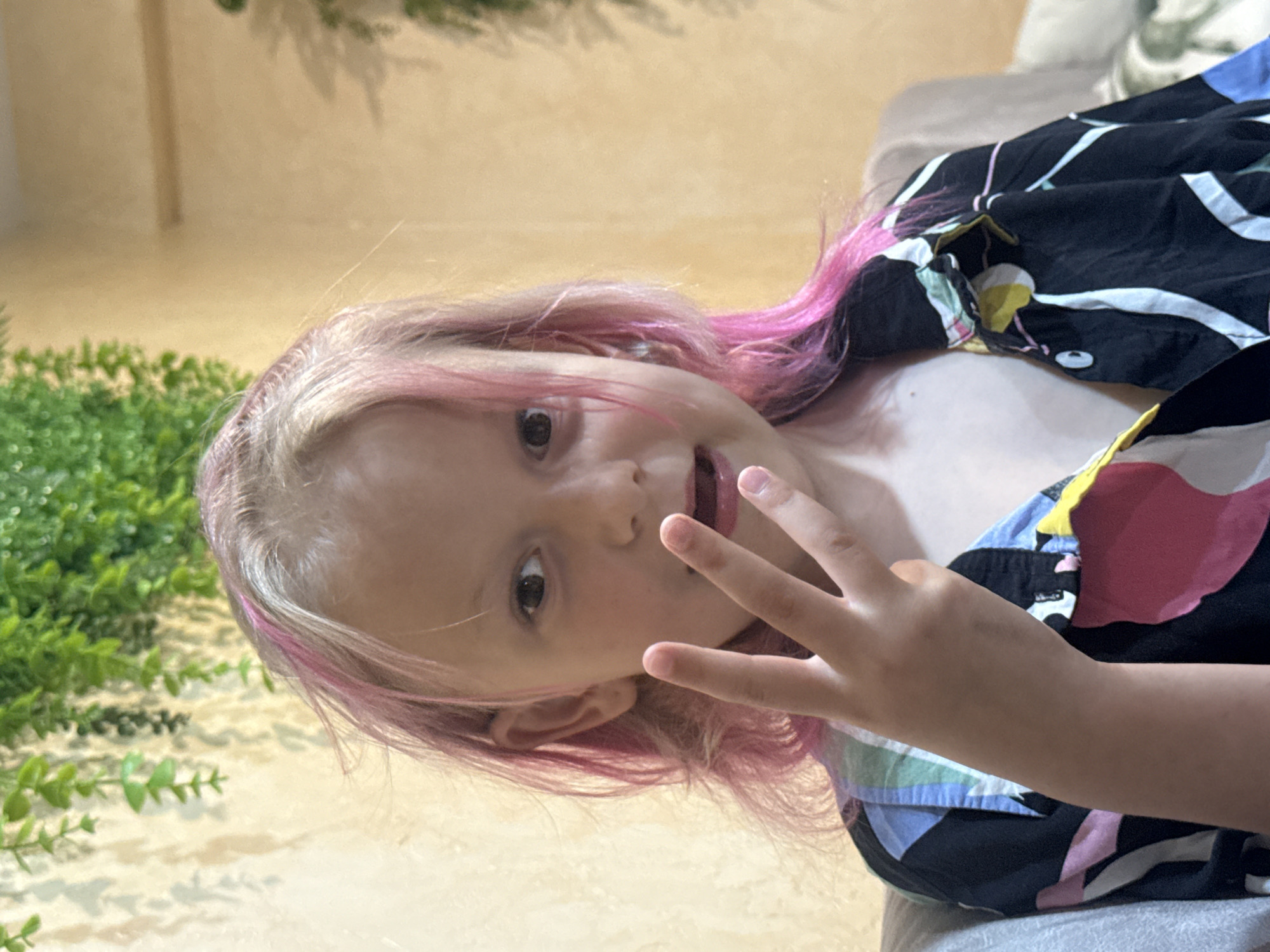 An eye-level shot captures a young person with light skin and blonde hair featuring prominent pink streaks, lying down and looking directly at the camera. They are making a peace sign with their right hand, holding up their index and middle fingers. A slight smile plays on their lips, and their dark eyes are open, engaging the viewer.
The subject is wearing a dark-colored shirt adorned with a vibrant, abstract pattern composed of various large, irregular shapes in shades of yellow, pink, blue, green, and white. The background is softly blurred. To the left side of the frame, behind the subject's head, there is a visible section of green foliage, possibly decorative plants, suggesting an indoor or semi-outdoor setting. The rest of the background consists of a light, warm-toned surface, possibly a wall or furniture. The subject appears to be resting on a light grey or off-white surface, such as a couch or bed. The overall impression is one of playful interaction in a relaxed environment. There is no visible text in the image.