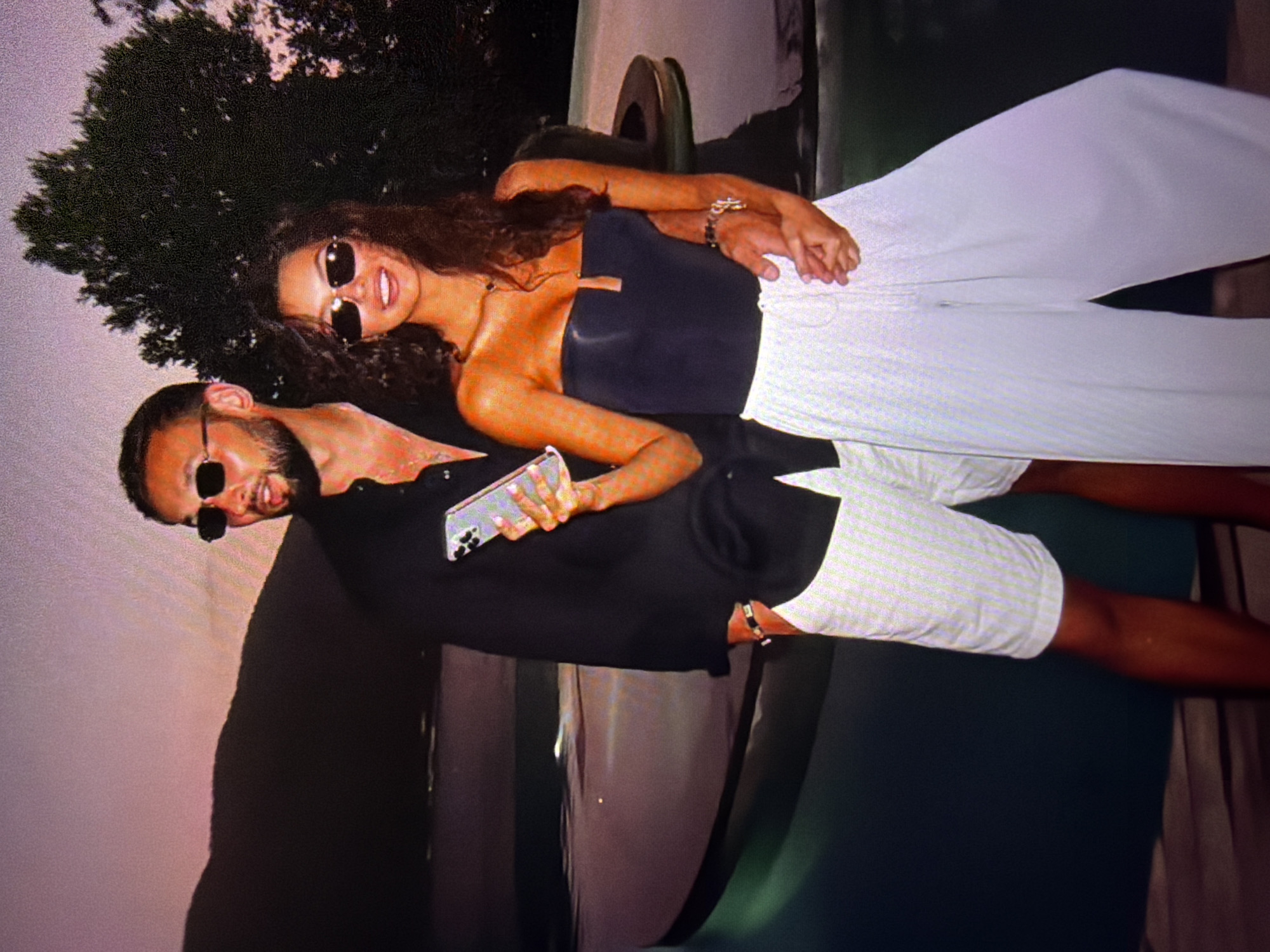 The image shows a couple standing by a pool during sunset. The man is wearing a black long-sleeved shirt, white shorts, and sunglasses. He is holding a smartphone in his left hand. The woman is wearing a black strapless top and wide-leg white pants, and sunglasses. She has her arm around the man's waist. The background features a dark body of water and a silhouette of a hill or mountain against a pink and purple sky. There is also a tree to the right of the couple. The scene suggests a luxurious vacation setting, potentially at a resort or a private residence. The presence of a pool and the sunset lighting indicate an outdoor evening setting.