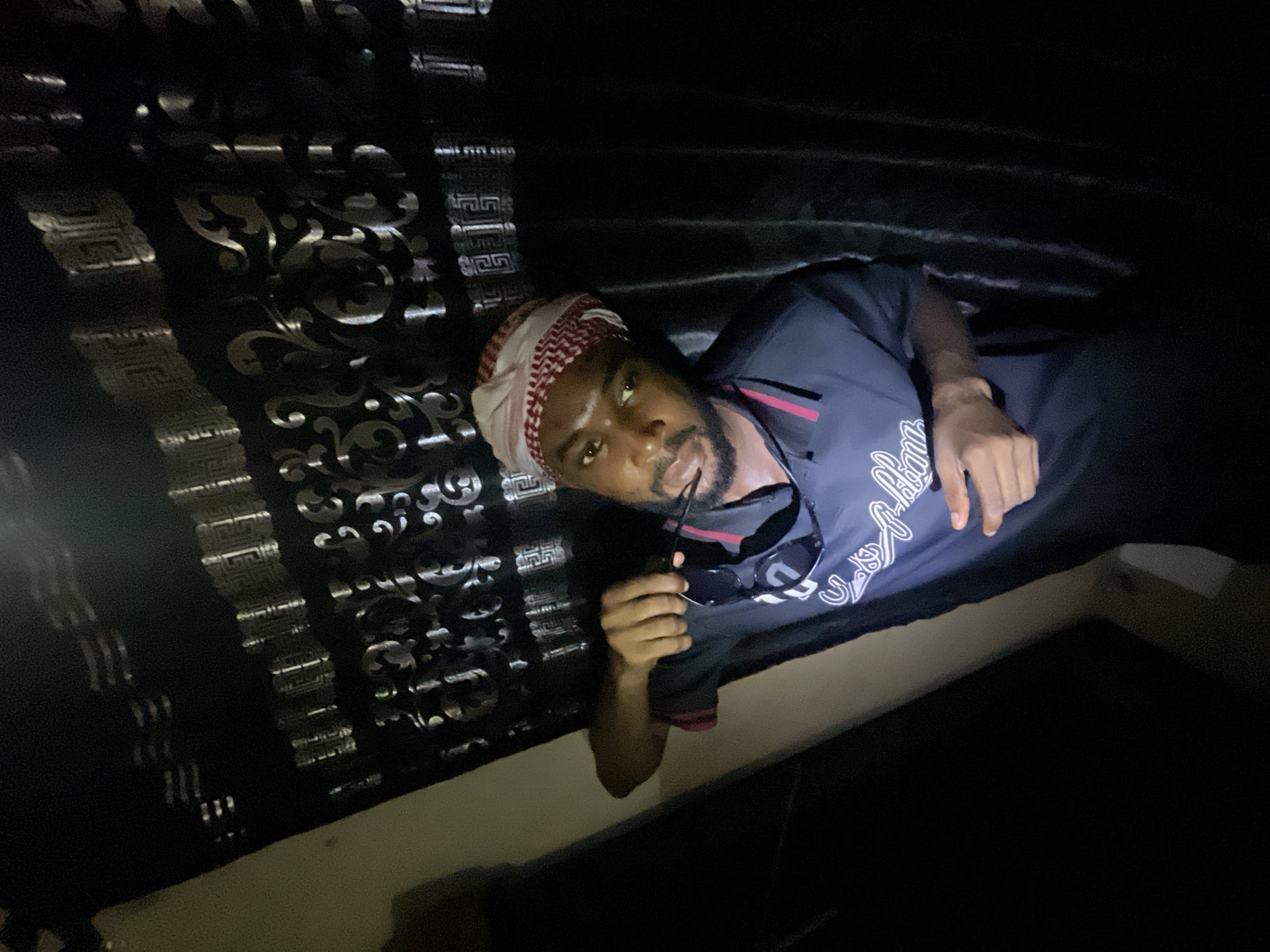 A man with dark skin and a beard is lying on a bed, wearing a kufi hat, holding a pair of sunglasses in his right hand. He is wearing a blue shirt with a white design. The bed has a black headboard with a silver patterned design. The location is unspecified, but the city is Lokoja, Nigeria. The lighting is dim, and the background is dark.