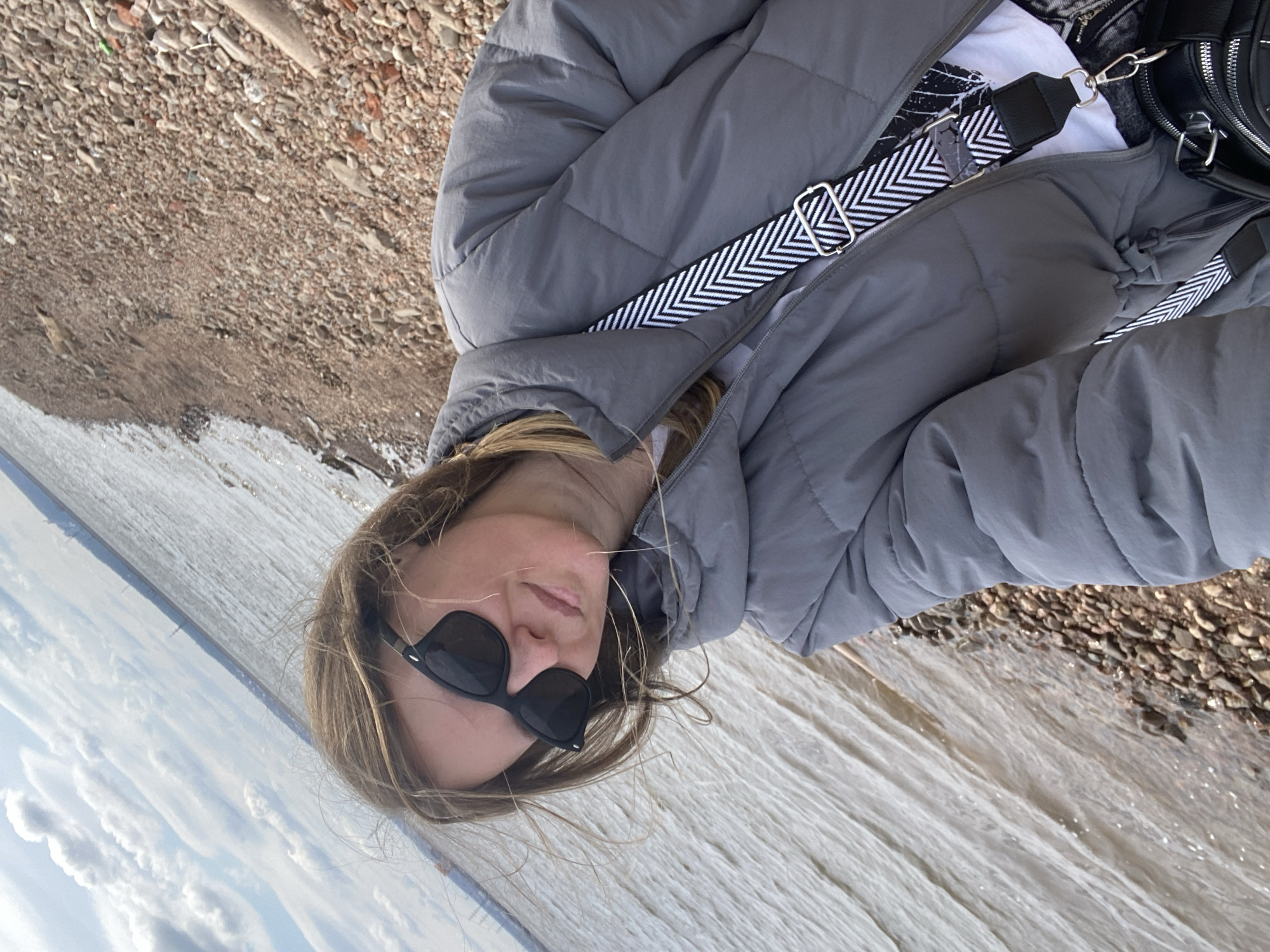 A person, likely a young woman, is captured in a close-up outdoor shot, posing for the camera. She has long, light brown hair, partially obscured by dark, square-framed sunglasses that cover her eyes. Her expression appears relaxed, with her lips slightly parted.
She is dressed in a warm, grey puffer jacket, which seems to be quilted, worn over a white t-shirt. A black shoulder bag with a prominent black and white chevron-patterned strap is visible across her body.
The setting appears to be a natural outdoor environment in Noviy, Russia. The person is positioned near a body of water, likely a river or lake, the surface of which shows ripples and reflections. The shore beside her is covered with various shades of brown and grey pebbles and small rocks. Above the water, a light blue sky with scattered white, fluffy clouds is visible, indicating a clear, possibly cool, daytime. There is no visible text in the image.