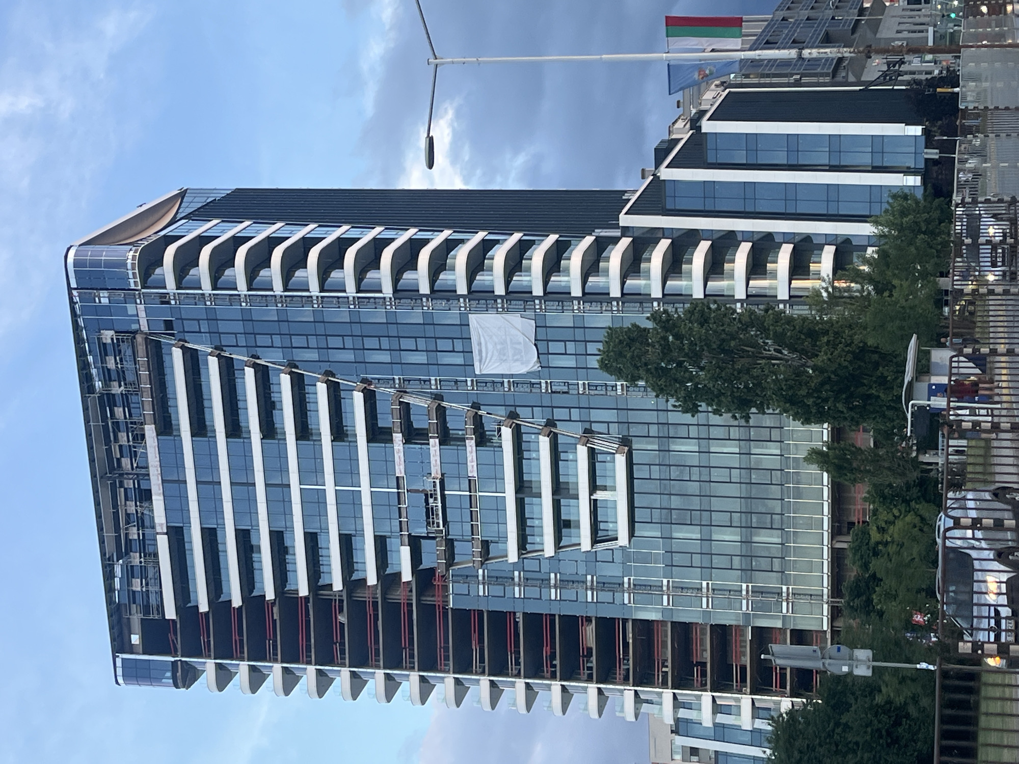 This is a low-angle, medium shot of a modern skyscraper under construction, photographed outdoors during the daytime. The building is predominantly glass and steel, with curved white balconies creating a distinct architectural feature on its upper floors. A large white banner, its text unreadable, is draped across a section of the building. Scaffolding is visible on several levels, indicating ongoing construction work. In the background, trees and other buildings can be seen. Two flags, one green, white, and red, and another blue, are flying from a flagpole at the top of the building. The sky is partly cloudy, with patches of blue and grey. The overall impression is one of urban development and industrial activity.