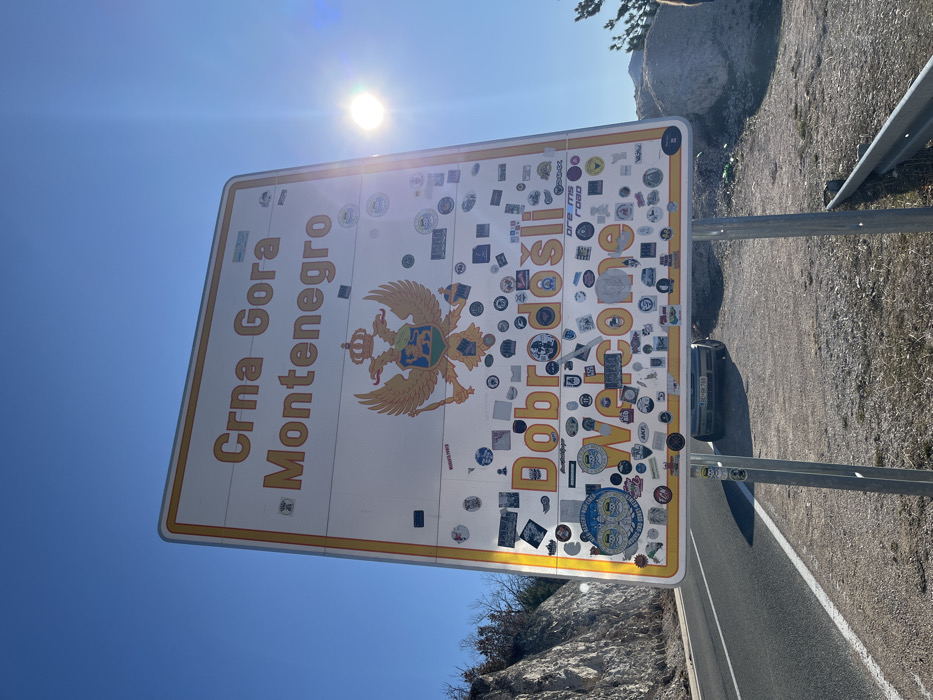 A large, rectangular white and yellow border sign, identifying entry into "Crna Gora Montenegro" with the Montenegrin coat of arms and the greeting "Dobrodošli Welcome," stands prominently at a roadside in Klobuk, Bosnia and Herzegovina. The sign's lower two-thirds are extensively covered with numerous small, multi-colored stickers. It is supported by two grey metal posts. To the left of the sign, an asphalt road with a solid white line runs alongside a rocky hillside with sparse, bare vegetation under a clear blue sky. To the right, a gravel and dirt shoulder features a partially visible light-colored car (license plate "DG BK 769") behind the sign's lower section, a metal guardrail, and scattered large rocks with sparse dry vegetation. The bright sun is visible in the upper right quadrant of the sky, causing lens flare. No individuals are visible.
