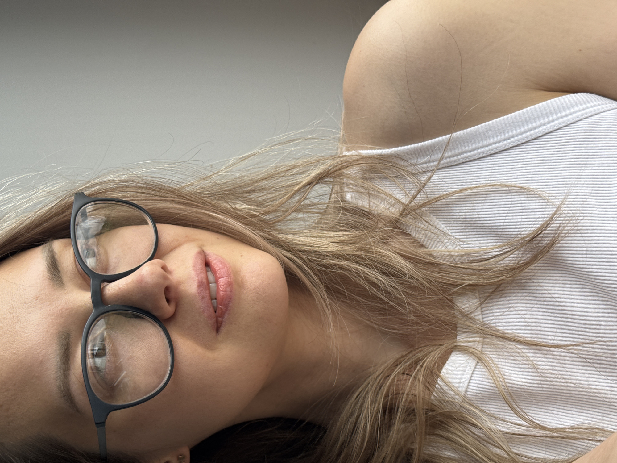 A woman with light brown hair is lying down, facing up and to the left. She is wearing black-rimmed glasses and a white striped tank top. The background appears to be a plain wall. There is no indication of the city of Chaikovsky, Russia in the image.