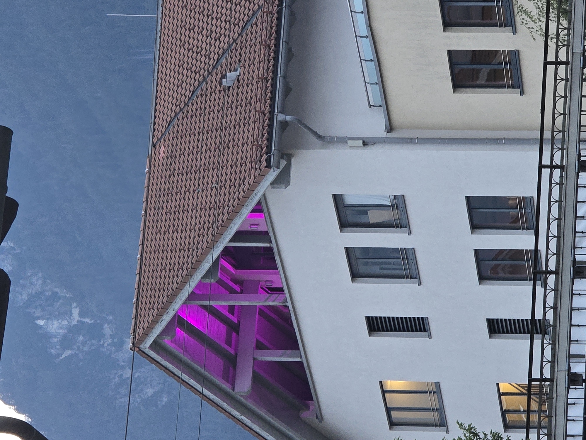 The image shows a close-up view of a building with a pitched roof covered in terracotta tiles. The attic space of the building is illuminated with bright pink lighting, visible through the gabled window. The building's facade is white and features several windows with dark frames. Some windows have visible blinds or shutters. A neighboring building, to the right and slightly lower, is painted a pale yellow and also has dark-framed windows. A metal railing with an arched design runs along the balconies of both buildings. In the background, the sky is a deep blue with some white clouds, and the silhouette of a mountainous region, likely the Alps, is visible. The overall scene appears to be set in the late afternoon or early evening, given the lighting conditions. The location is identified as Garmisch-Partenkirchen, Germany. There are no people or animals clearly visible in the image.