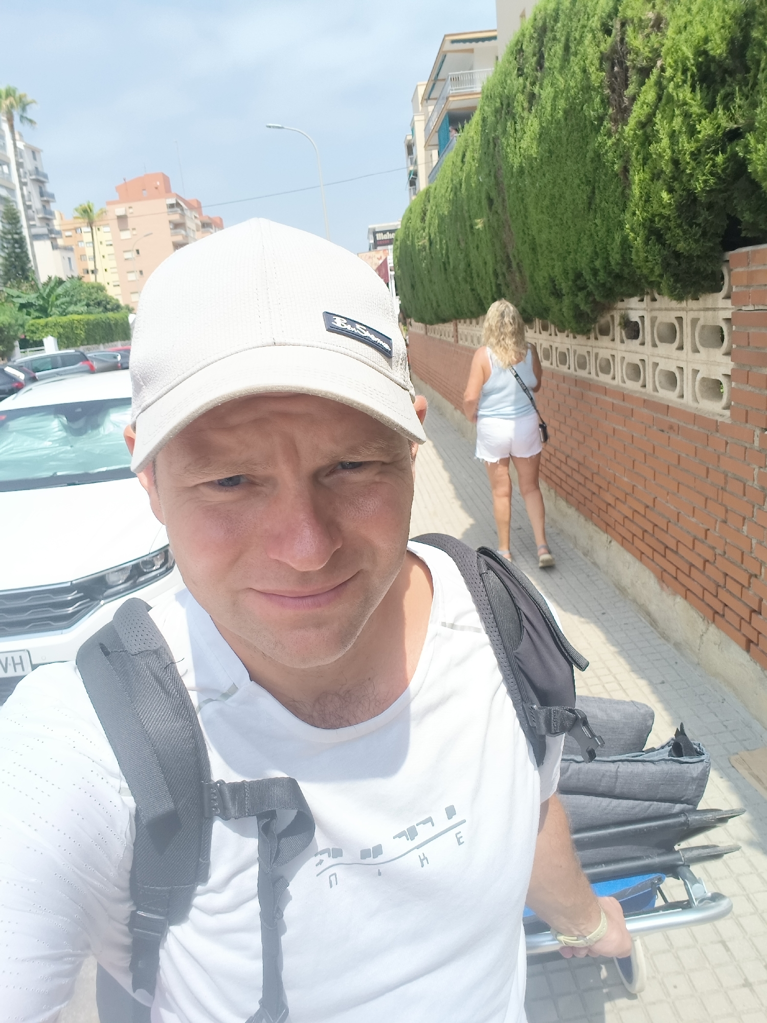 A man wearing a white t-shirt and a light-colored baseball cap is taking a selfie on a sunny day, holding the camera with his right hand. He is wearing a backpack. In the background, a woman with blonde hair wearing a light blue tank top and white shorts walks away from the camera. She is wearing a crossbody bag. They are on a paved sidewalk next to a brick wall and a row of manicured green bushes. Buildings and palm trees are visible in the distance, suggesting an urban or semi-urban environment. A white car is parked on the left side of the frame. The license plate is partially visible and reads "WH". The overall scene suggests a casual stroll in a warm climate, possibly in the city of Gandia, Spain, given the architectural style and vegetation. The man is also pulling a wheeled cart carrying stacked gray items, possibly luggage.