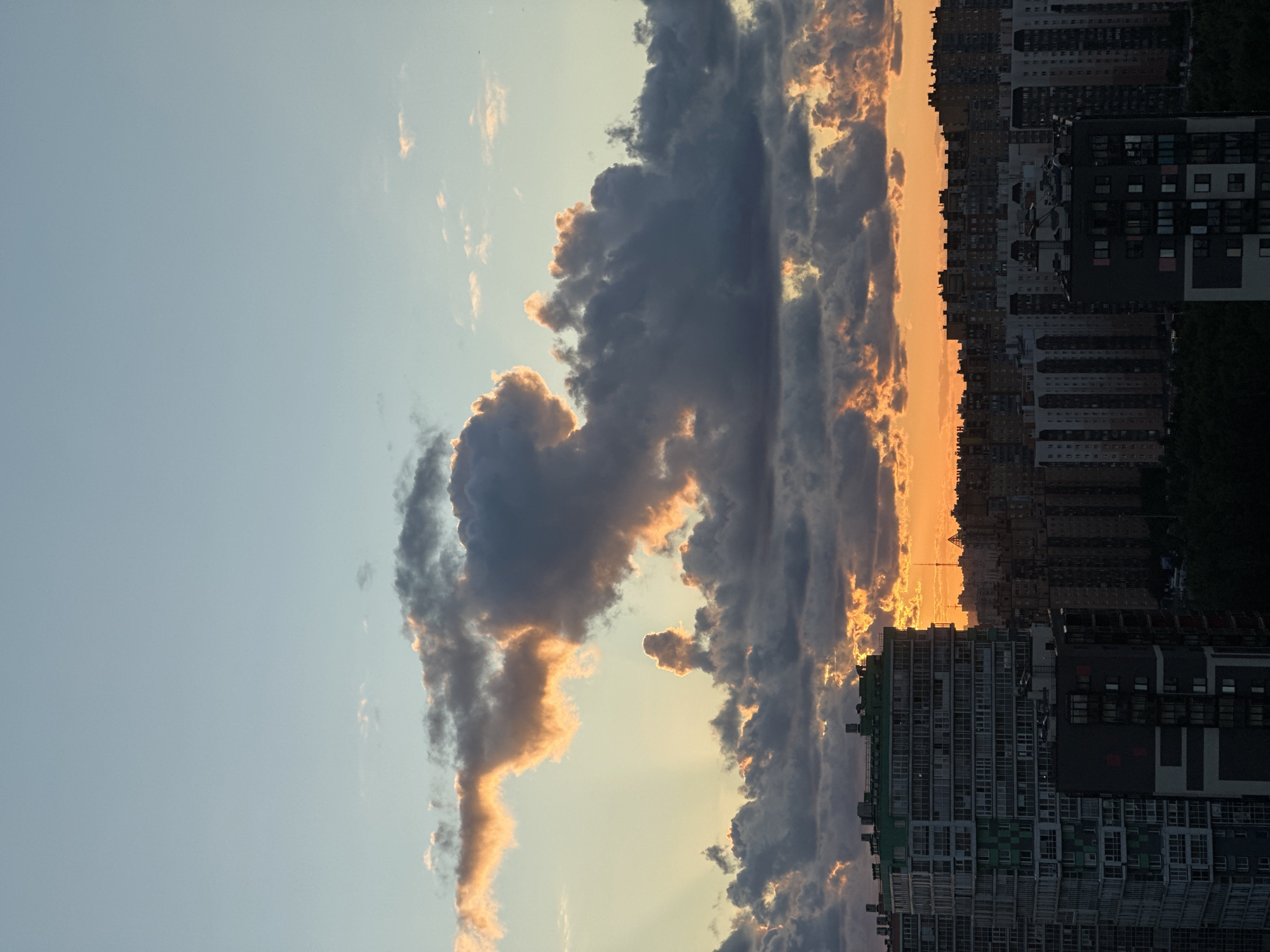 A vertical, medium shot captures a city skyline at sunset. The sky is a gradient of pale blue at the top, transitioning to lighter shades of blue and yellow, and finally to a vibrant orange near the horizon. Large, dark gray clouds are illuminated by the setting sun, creating bright orange and yellow highlights along their edges. The sun's rays pierce through gaps in the clouds, casting beams of light downwards.
In the foreground, the tops of several multi-story apartment buildings are visible. The building on the left is tall and modern, with a green section on its upper floors and many windows. To the right of this, a darker, shorter building is positioned. Further into the background, a dense cluster of residential buildings stretches across the horizon, their silhouettes dark against the bright sunset. The overall impression is of an urban environment during dusk, with dramatic cloud formations and the warm glow of the setting sun. The location is identified as Dnp Ryzhiki, Russia. There are no people or discernible actions depicted in the image.