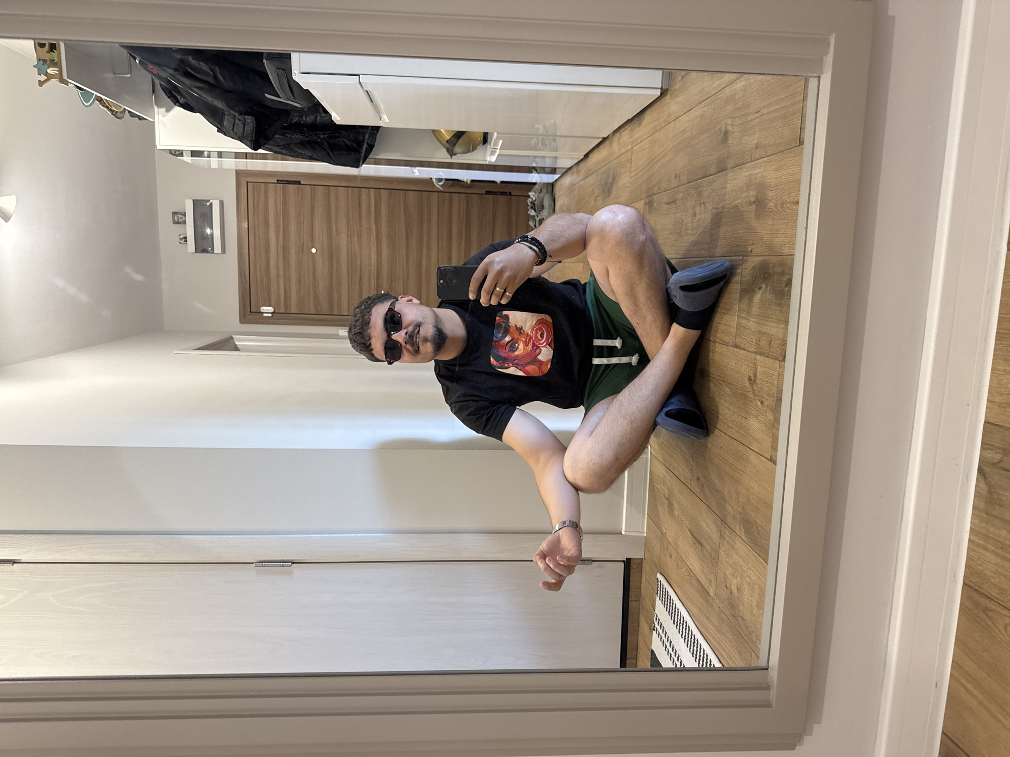 A mirror image shows a man squatting, taking a photo with his mobile phone. He's wearing sunglasses, a black t-shirt with a colorful graphic, green shorts with white stripes, and blue slides. He has multiple bracelets on his left wrist and a watch on his right. The setting is a hallway with light wood flooring. In the background, there's a light-colored door and what appears to be closet or storage space with a black jacket hanging. A small rug is visible at the edge of the reflection. The city and specific location are unknown.