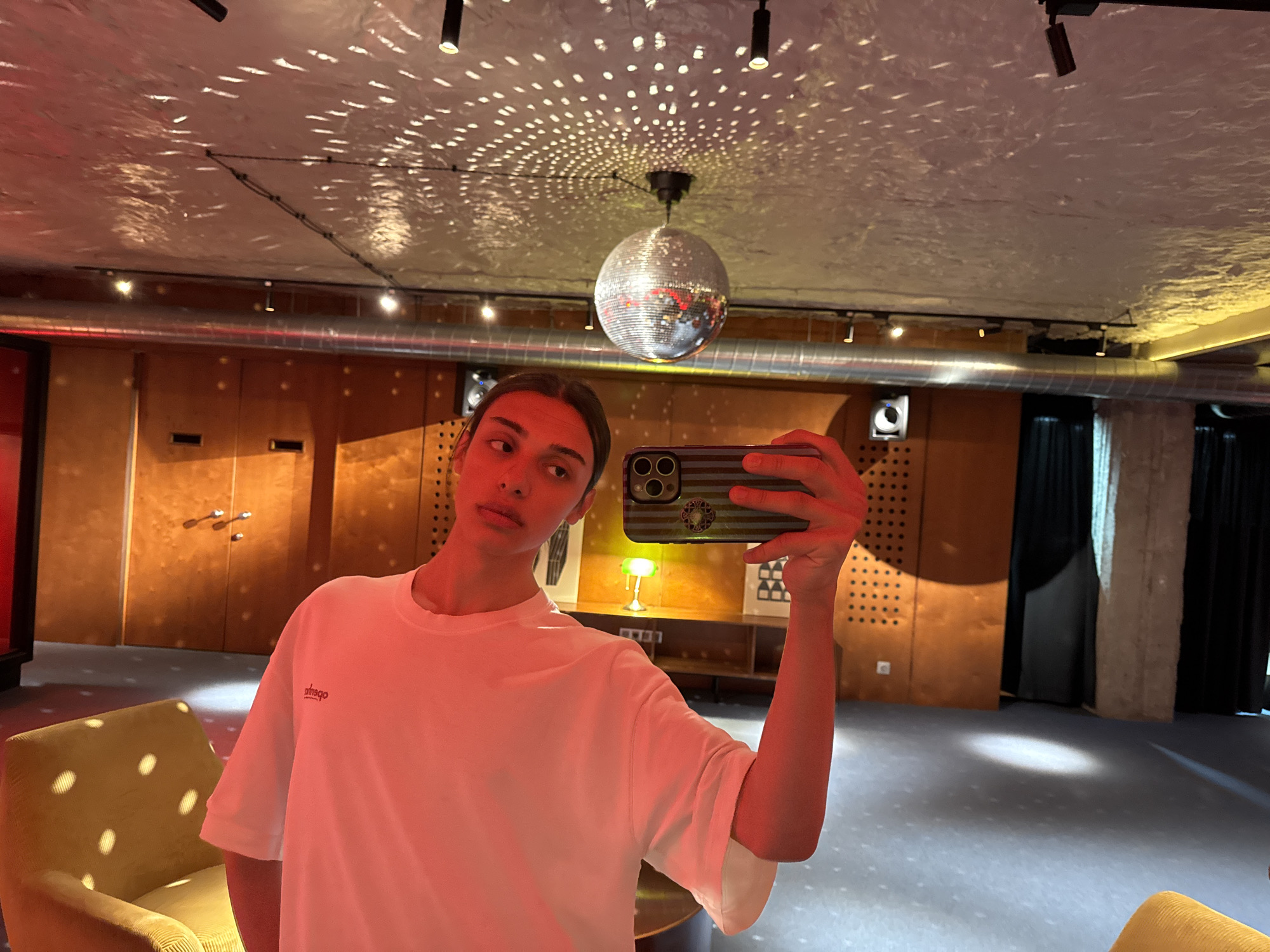 A young adult is taking a selfie in a room with retro-style décor. The person is wearing a white t-shirt with small dark lettering on the upper left chest. The phone they are using has a dark-colored case with horizontal stripes and circular design elements. Above the person's head hangs a disco ball, and the ceiling is covered with reflective light patterns. The room features wood-paneled walls, a yellow upholstered chair, a silver air duct running across the ceiling, and a concrete support pillar visible in the background. There is a lamp and artwork on a shelf behind the person. The floor is covered in blue carpet with light-colored spots, possibly reflective. The room appears to be located in Yerevan, Armenia, based on architectural styles and furnishings.