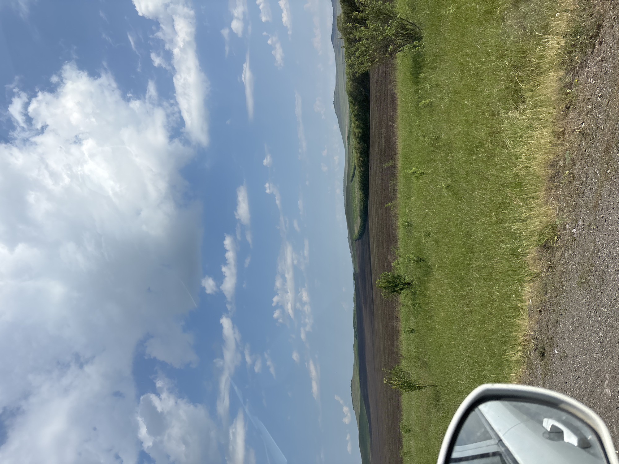 An outdoor, eye-level, full shot captures a landscape scene from a vehicle's side mirror. The mirror, reflecting the car's white door handle, is in the lower-left corner of the frame. Beyond the mirror, a gravel road leads into a field of green grass, dotted with a few small trees. Further back, a dark brown field sits below a line of green trees, which in turn is beneath rolling green hills under a bright blue sky filled with white, fluffy clouds. The scene appears to be of the fields on the outskirts of the city of Borodino, Russia.