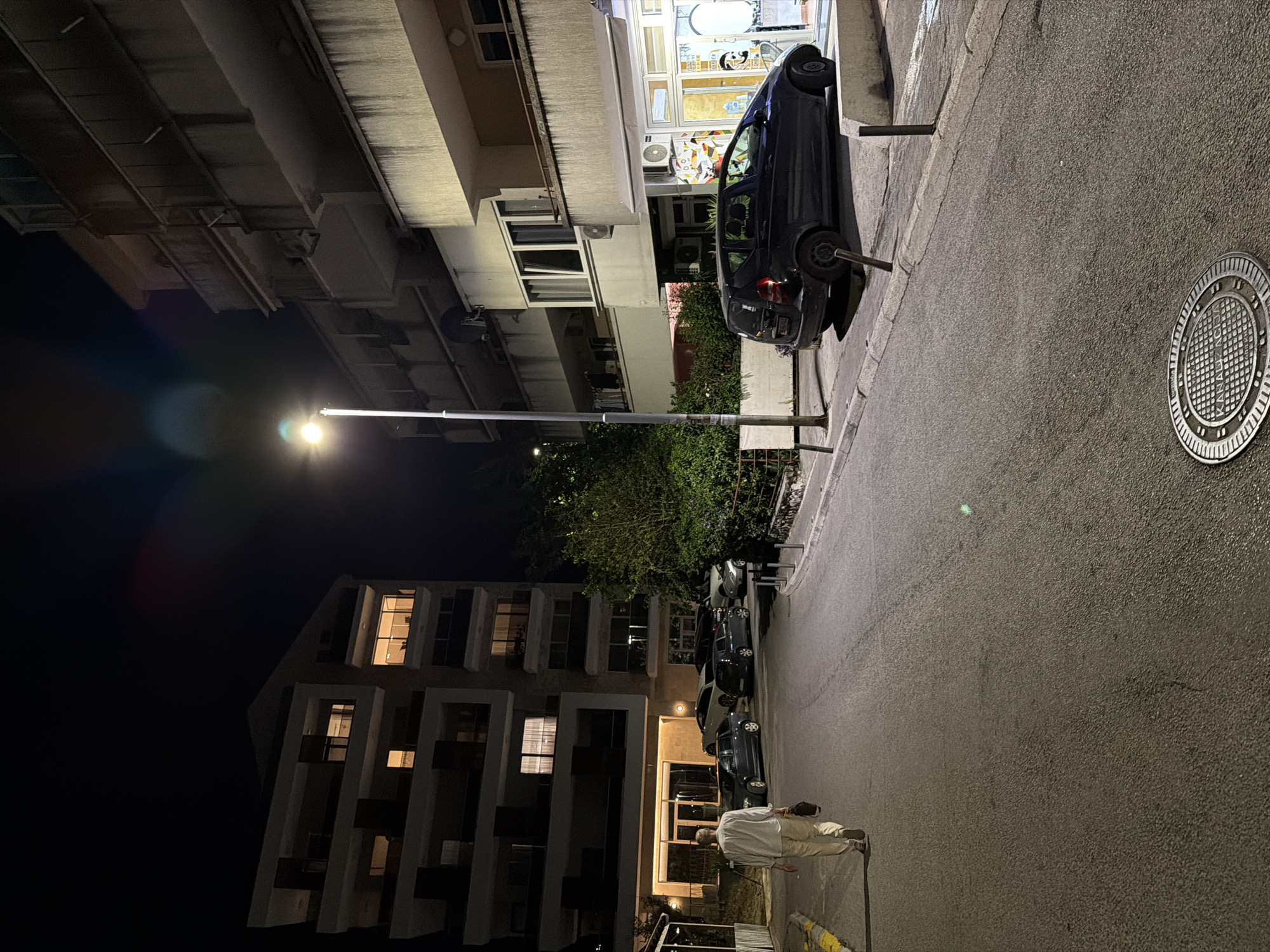 The image depicts a nighttime scene on a street in Budva, Montenegro. A man, wearing a light-colored shirt and pants, walks away from the camera down the asphalt street, carrying a dark bag over his shoulder. On the left side of the street, a multi-story building with lit windows is visible. On the right side, another multi-story building houses a business with a lighted storefront. Parked cars are visible along the street in front of both buildings. A street lamp illuminates the scene. The environment appears to be a residential or mixed-use urban area.