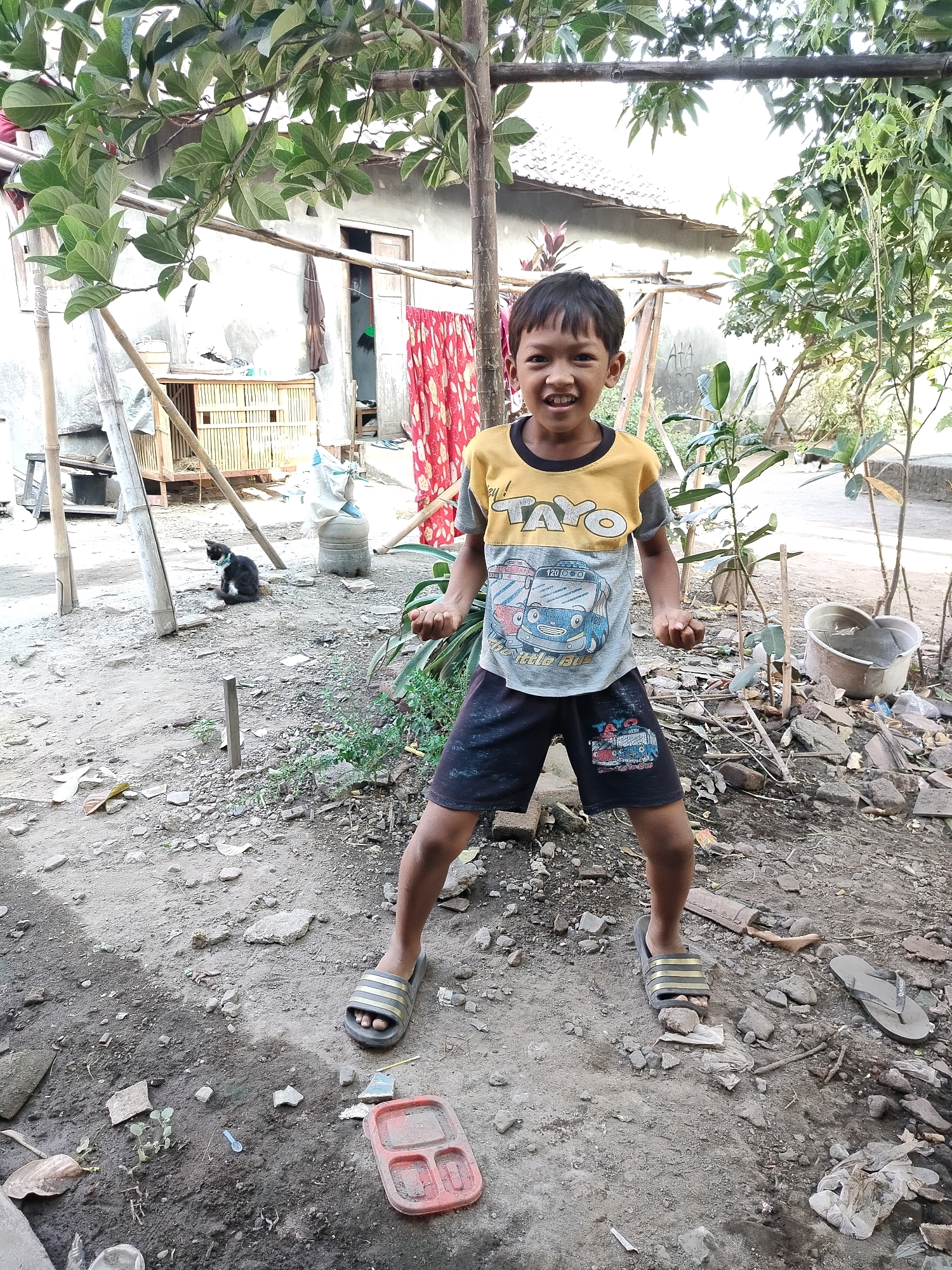 A young boy is standing in an outdoor area, possibly a backyard, in Beloh, Indonesia. He is smiling and looking directly at the camera. He is wearing a yellow and gray t-shirt with cartoon bus graphics and dark shorts. He has on dark-colored sandals. The ground is dirt and gravel, with some scattered debris, including rocks, leaves, and a red plastic container. In the background, there is a house with a gray wall and a tiled roof. There are also some plants and trees, a wooden structure, and a black cat sitting on the ground. The overall scene suggests a casual, everyday environment in a residential area.