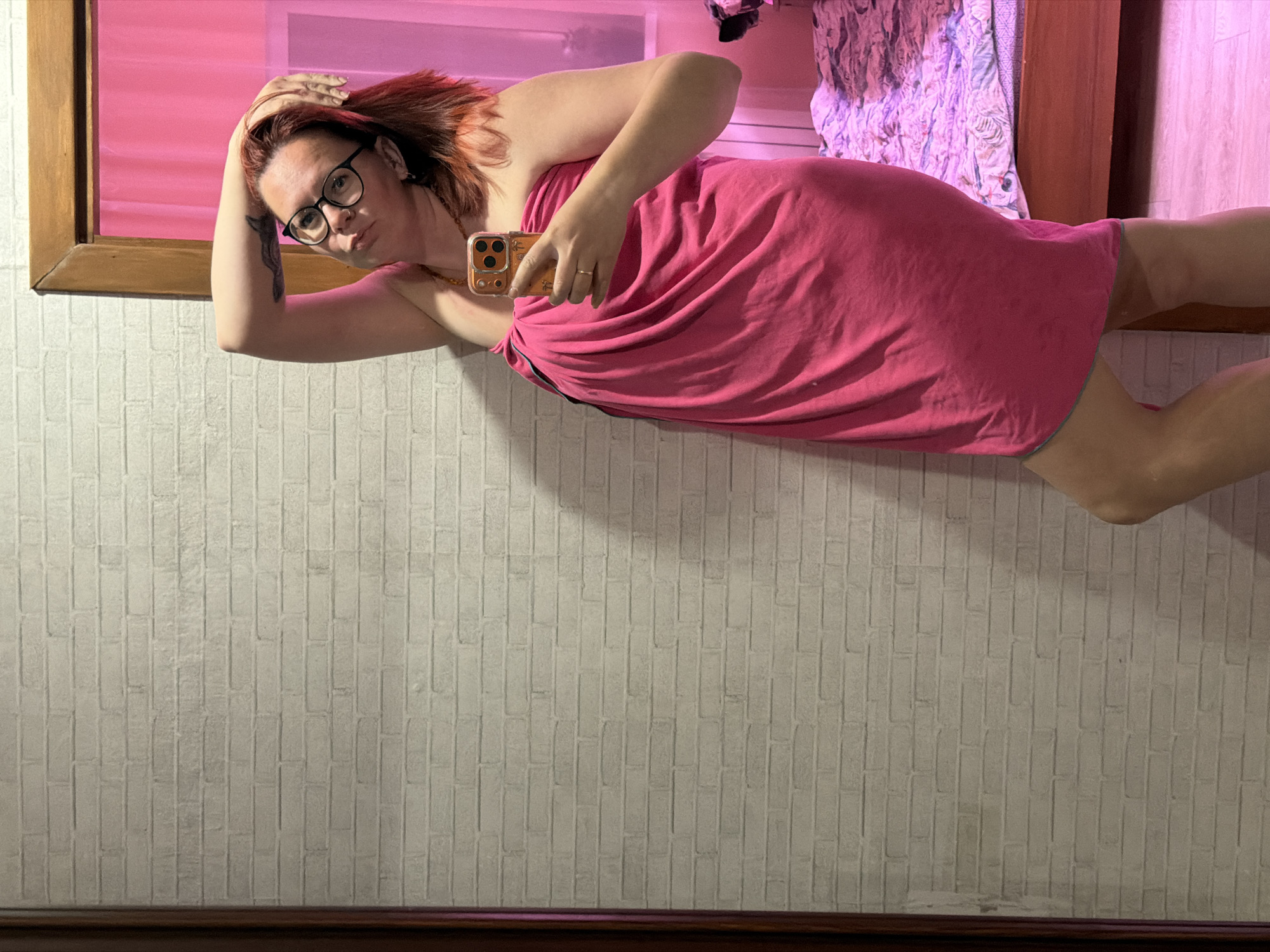 The image shows a woman taking a mirror selfie in an indoor setting, likely a bedroom or dressing area, in Chaikovsky, Russia. She is positioned to face the camera, holding her smartphone with a gold-colored case in her right hand. Her left hand is raised and resting on her head or in her reddish-brown hair.
The woman is wearing a strapless pink dress or a towel wrap, and black-rimmed eyeglasses. She has a slight pout on her face and is looking directly towards the camera. A small tattoo is visible on her left upper arm, and she wears a ring on her left ring finger. The mirror, framed with light-colored wood, reflects a strong pink and purple hue, especially in the upper section, where a patterned fabric, possibly a bedspread or curtain, is also visible. The wall behind her and below the mirror is covered in light-colored wallpaper with a textured, brick-like pattern. A dark wooden element, possibly a headboard or baseboard, runs along the very bottom of the frame. The lighting suggests an interior space, potentially with artificial lighting contributing to the prominent pink coloration in the mirror's reflection.
