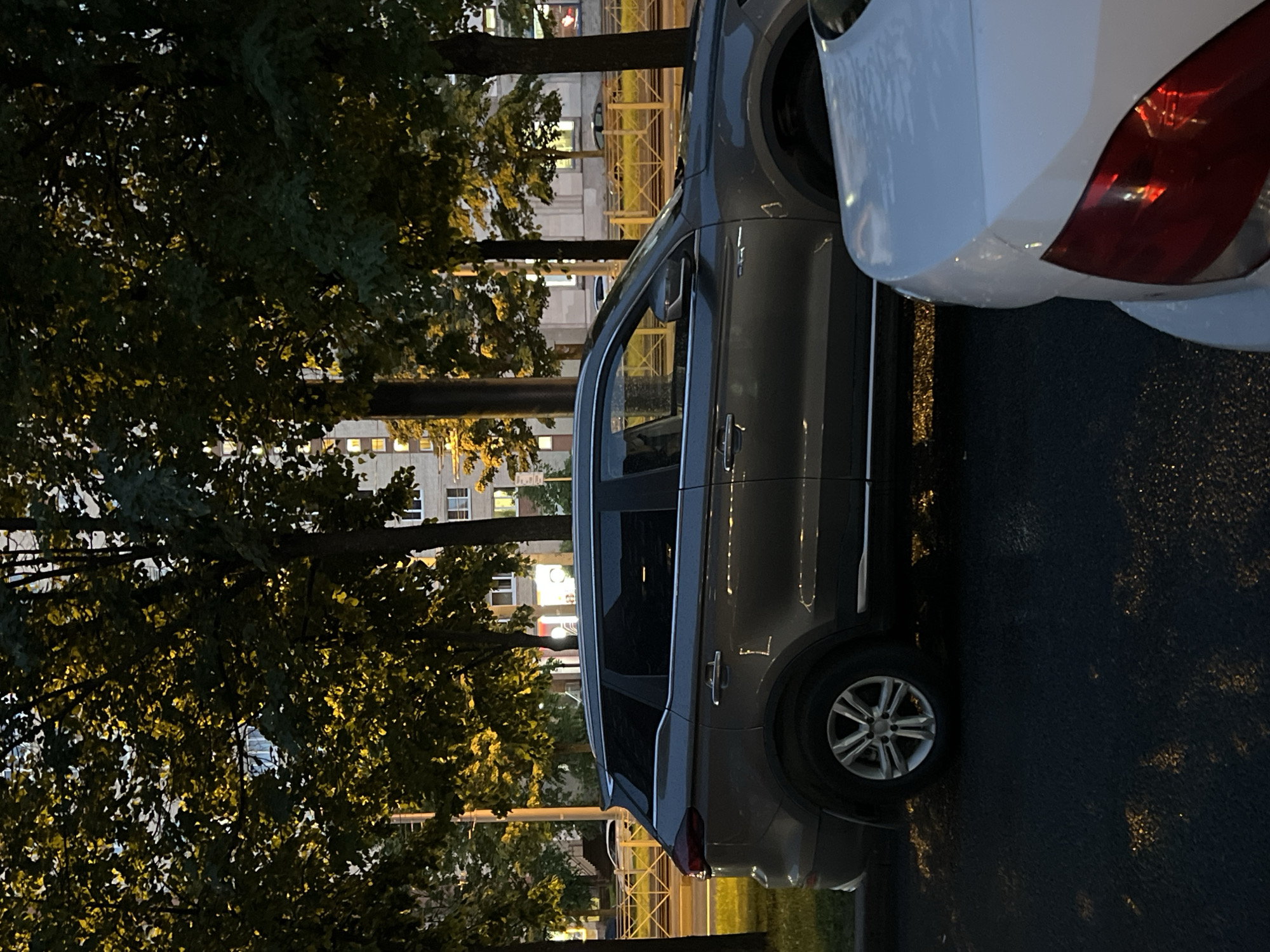 The image shows a gray SUV parked on an asphalt surface, partially obscured by trees with dense foliage. The trees are illuminated from behind, suggesting the presence of streetlights or building lights. In the background, a multi-story building with illuminated windows is visible, indicating an urban or suburban setting.
A portion of a white car, likely another SUV or a similar vehicle, is visible on the right side of the image, parked next to the gray SUV. The white car's rear light is visible, and its body appears to be wet, possibly due to rain or recent washing.
The time of day appears to be evening or night, given the artificial lighting and the dark sky. The overall scene suggests a quiet moment, possibly after sunset, in Sinyavino, Russia. There are no people or specific activities depicted. The focus is on the parked vehicles and the surrounding environment. The mood is calm and uneventful. No visible text is present in the image.