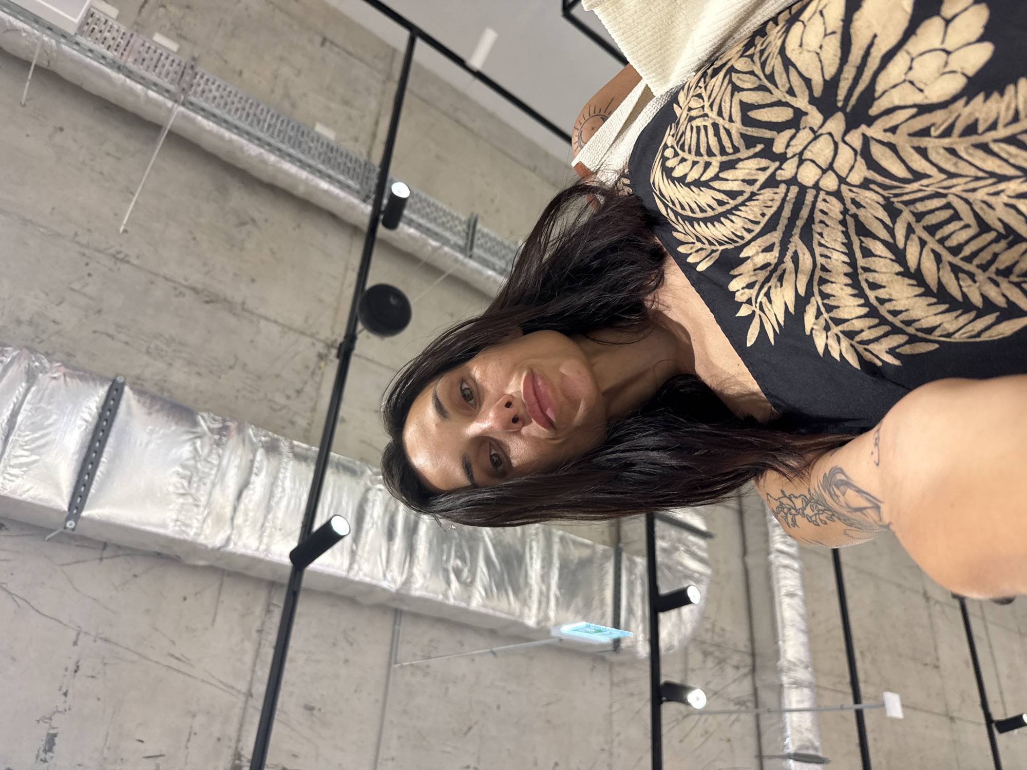 The image captures a woman posing for a photo against an industrial-style interior background. She has long, dark hair, dark eyes, and full lips, looking directly at the camera with a neutral expression and a slight pout. She is wearing a black top or dress with a striking gold or tan botanical pattern, resembling a large stylized flower or plant.
Visible tattoos include a design featuring a sun and crescent moon on her right upper arm/shoulder, and a partially visible, potentially floral or abstract, tattoo on her left arm.
The background features a textured concrete wall. Exposed silver ventilation ducts, possibly wrapped in insulation, are prominent, running horizontally across the upper and lower parts of the wall. Black track lighting fixtures with bright spotlights illuminate the space, mounted on vertical and horizontal black poles. A small, light-blue rectangular object, possibly a suspended display or sign, is visible in the lower center background. The setting suggests a modern, minimalist interior, characteristic of a studio, gallery, or commercial space in Pleven, Bulgaria. No discernible text is visible.