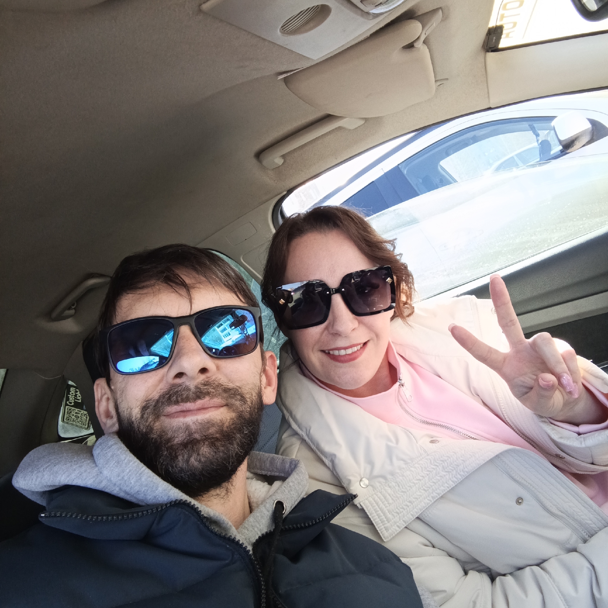 A man and a woman are in a car, likely taking a selfie. The man has dark hair and a beard, wearing blue sunglasses and a blue jacket with a gray hoodie underneath. The woman has dark hair, large black sunglasses, a pink shirt, and a white jacket. She is holding up two fingers in a peace sign. The interior of the car is visible, with a light-colored roof and dashboard. A sticker is visible on the driver's side window. It cannot be determined whether this scene is in the city of Марковский, Russia.
