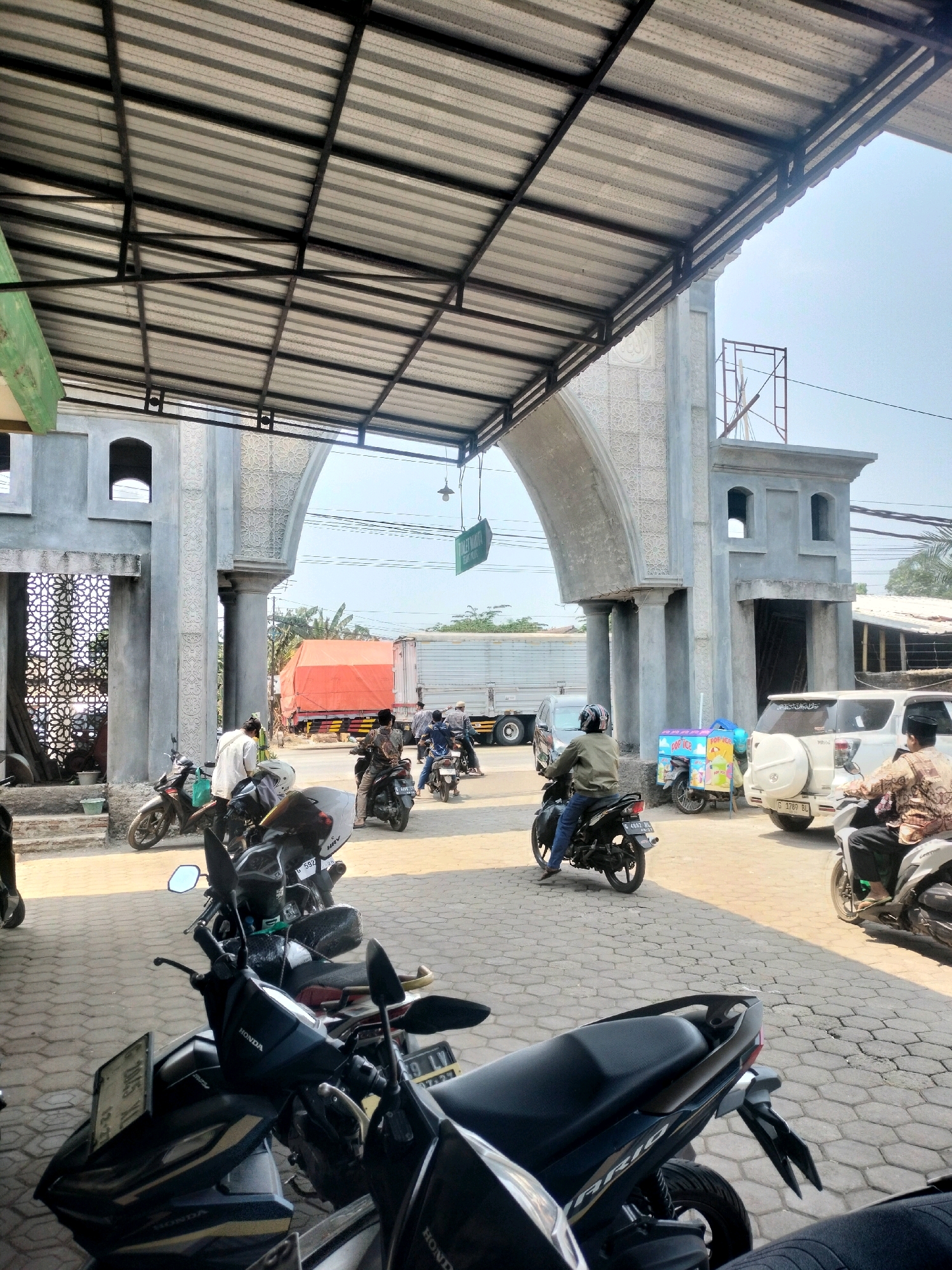 The image captures a street scene in Kutosari, Indonesia, under a covered walkway supported by metal beams and a corrugated metal roof. An arched gateway with Islamic-inspired decorative patterns is prominent in the background, leading to buildings under construction or renovation. Several motorcycles are parked and in motion on a paved ground.
In the foreground, a black motorcycle is parked, its license plate partially visible. Behind it, other motorcycles, some with riders, are present. To the right, a white SUV is parked, and a person on a motorcycle is moving away from it. Further back, a person on a motorcycle is approaching the archway. An ice cream cart is also visible near the white SUV.
In the distance, a large orange-covered truck is parked alongside a white truck. The sky is clear and bright. The overall impression is a busy street in a developing area, likely near a religious or community center given the architectural style.