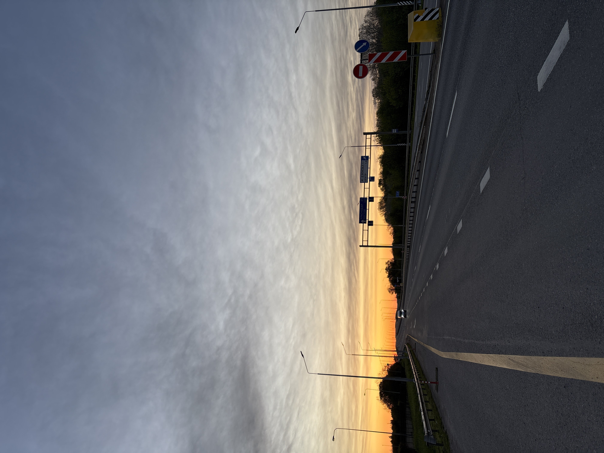 The image depicts a multi-lane highway or motorway during sunset or dusk, set in the context of Gelendzhik, Russia. The asphalt road, marked with white dashed lane lines and a solid yellow line on the shoulder, stretches from the foreground towards the distant horizon. Metal crash barriers line the road's edge.
The sky transitions from a cool blue-grey in the upper left to vibrant orange and yellow hues near the horizon on the right, illuminated by the setting sun. Streaky, elongated clouds are visible across the sky. Numerous streetlights recede into the distance along the highway.
Overhead gantries and poles display various road signs. Visible Cyrillic text includes "ЖД ВОКЗАЛ" (Railway Station) and "ЖЕЛЕЗНОДОРОЖНЫЙ" with distances "36" and "15" (likely kilometers), "КРАСНОДАР 45" (Krasnodar, 45 km away), and "СВХ" (an abbreviation likely for a bypass or express road). Other signs include a red circular 'no entry' or 'forbidden' symbol and a blue circular 'mandatory direction' sign. A possible route marker "11C" is also present. Trees and dense foliage create a dark silhouette against the warm sky along the roadside. A lone white car is visible in the distance on the highway, and a yellow and black striped safety barrier or sign is seen near the road's right edge. The scene evokes a tranquil sense of evening travel.