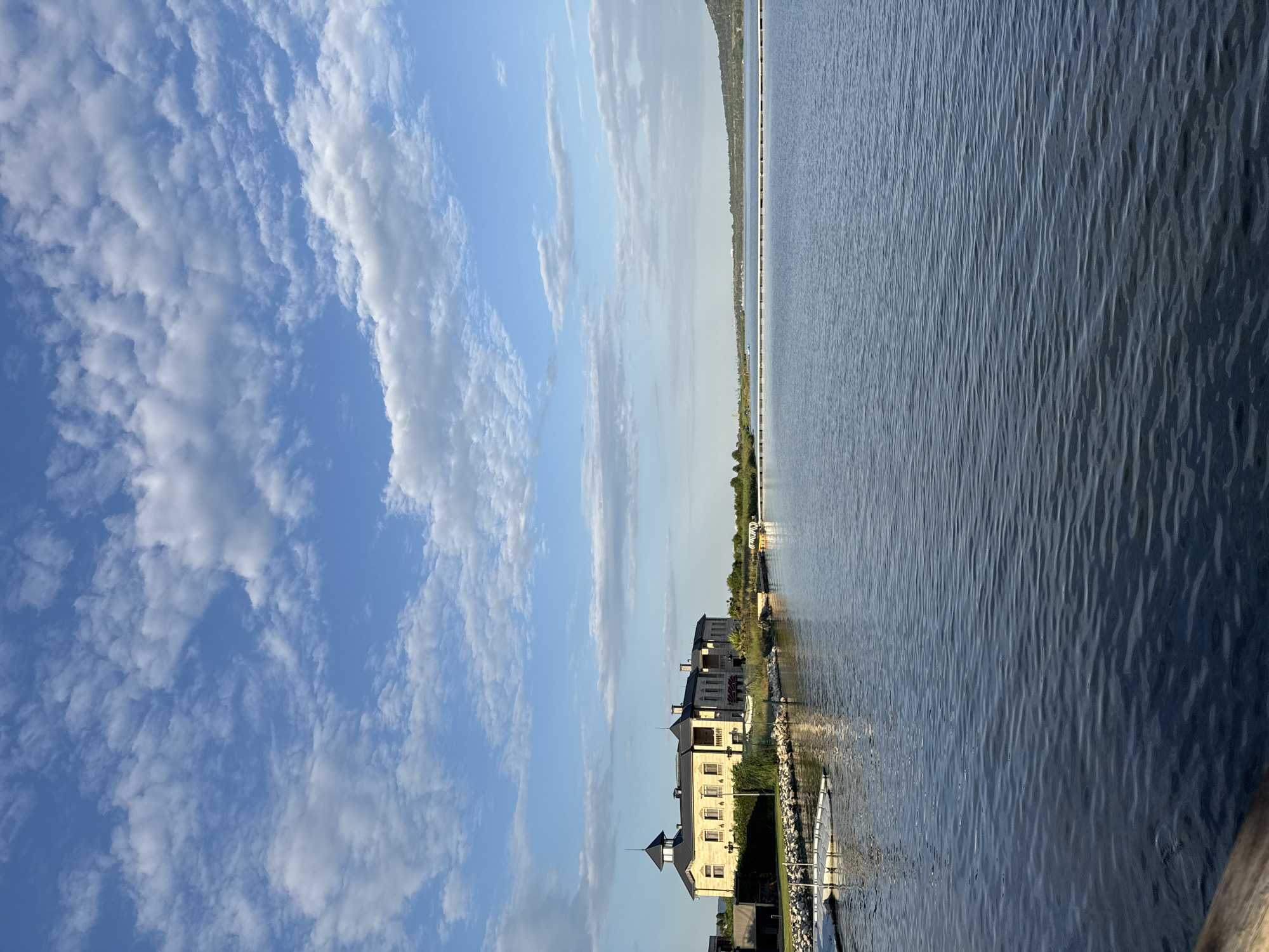 The image depicts a tranquil waterfront scene with a large, light-colored building in the background, possibly a hotel or a private residence, situated on the shore of a calm body of water. The building features multiple stories, dark-tiled roofs with turrets, and numerous windows. In the foreground, the water is dark blue and slightly rippled, reflecting the sky. Along the left bank, there is a dock with white floating platforms, suggesting a marina or a recreational area. Beyond the building, a distant shoreline is visible with trees and a faint outline of hills. The sky is a clear blue with scattered white clouds, indicative of fair weather. The overall impression is one of serenity and natural beauty. The location is identified as Казашко, Bulgaria.