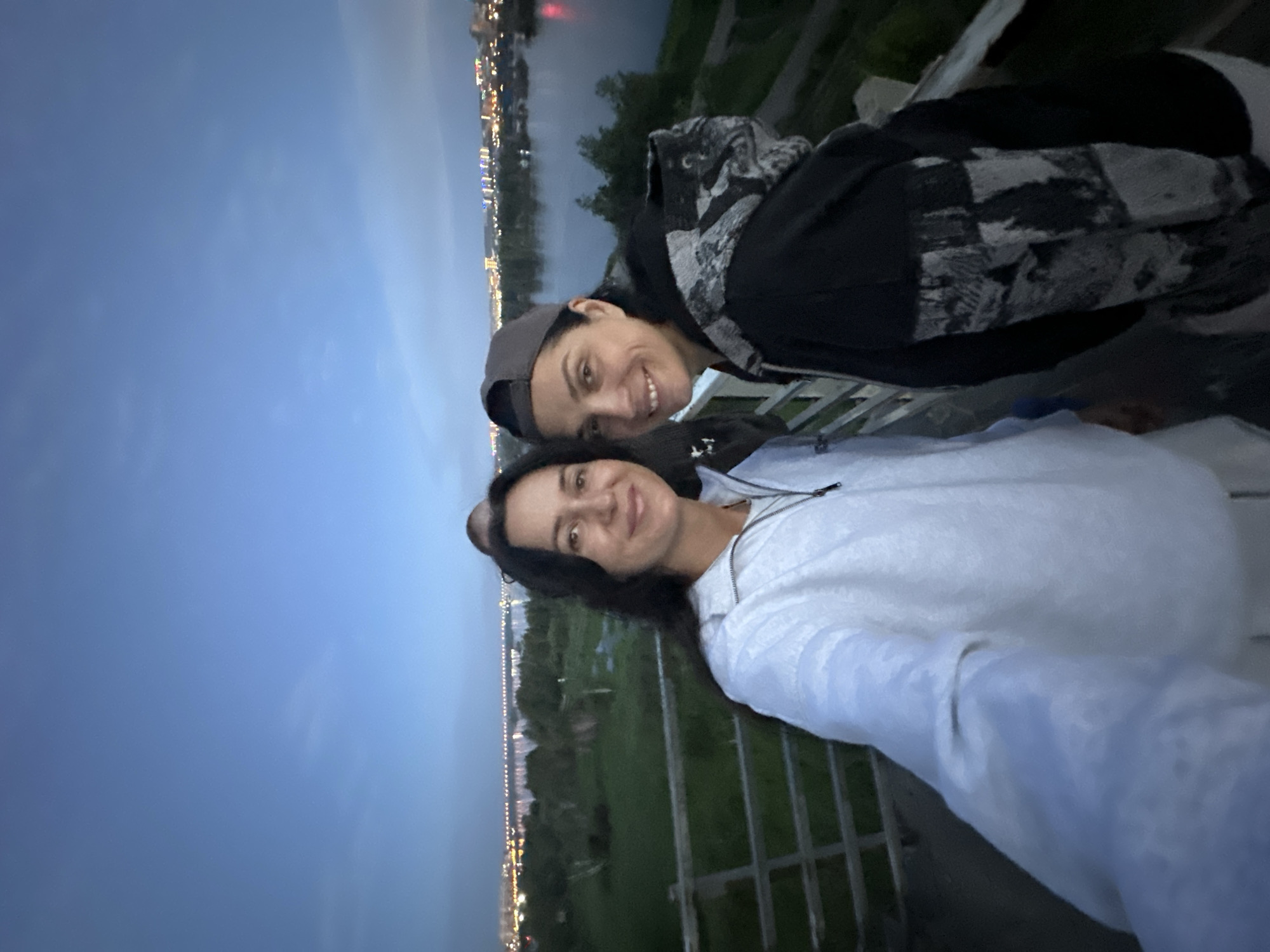 The image shows two women taking a selfie on a balcony or viewing platform overlooking a body of water, likely the Angara River, in Irkutsk, Russia. The cityscape with illuminated buildings and a bridge can be seen in the background. The sky is a deep blue, indicating it is dusk. The woman on the left is wearing a white hooded jacket, and the woman on the right is wearing a black hoodie and a baseball cap. There are faint lights and trees visible along the riverbank. A railing is present in the foreground.