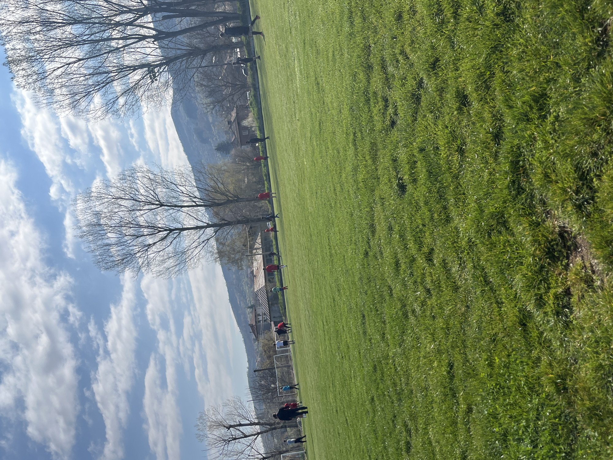 The image depicts a grassy field on a sunny day in Unknown, Unknown. Several people are dispersed across the field, appearing to engage in various activities. Trees line the edge of the field, and a mountain is visible in the background. The sky is blue with scattered clouds. A building is visible behind the trees. A soccer goal can be seen in the lower part of the image.