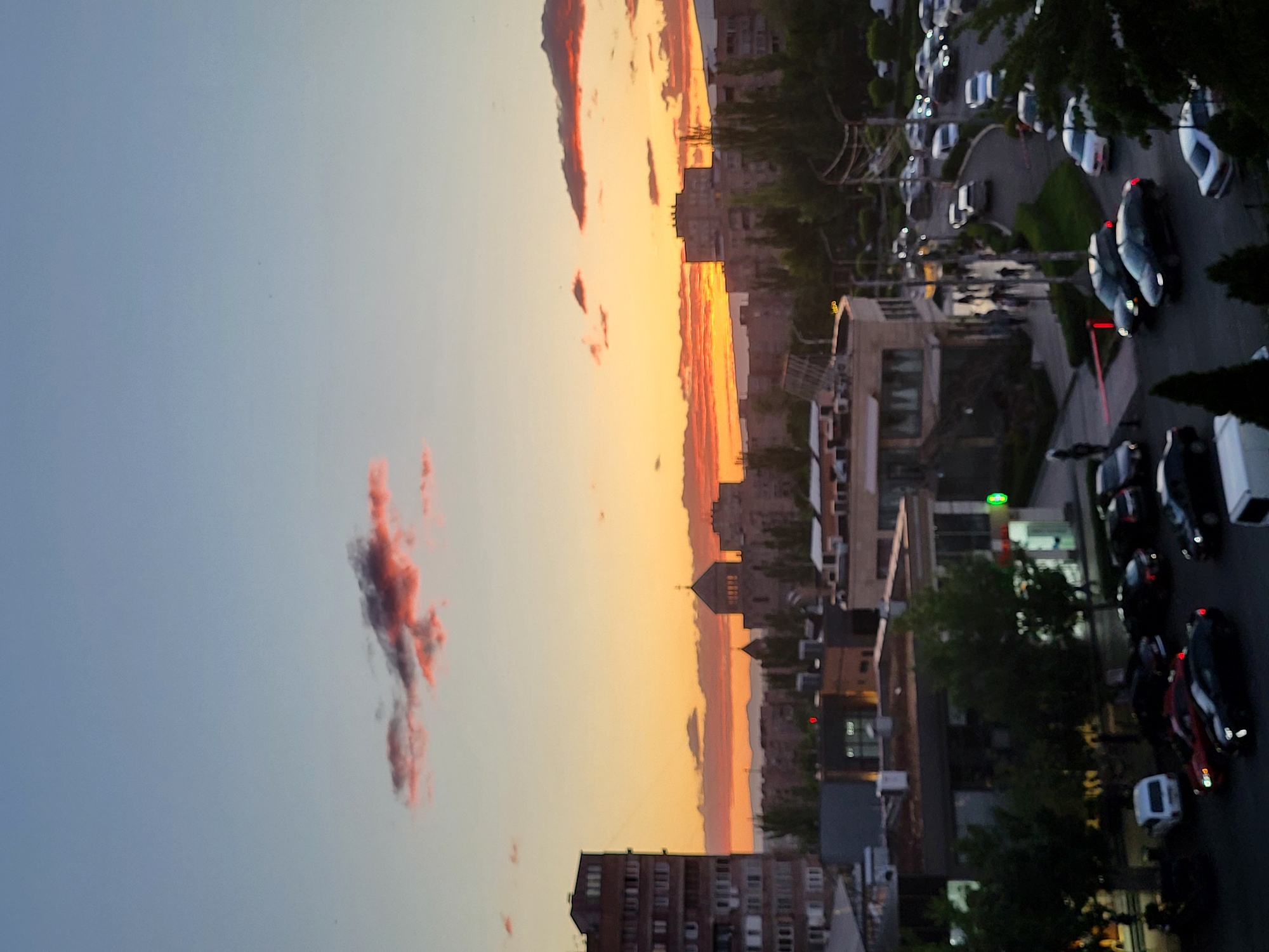 This media file captures an urban landscape in Yerevan, Armenia, during either sunset or sunrise, indicated by the striking sky colors. The image, though rotated, depicts a sky transitioning from a soft light blue at the top to a vibrant orange and yellow hue near the horizon. Several distinct, dark reddish clouds are scattered across this colorful expanse.
Below the horizon, the cityscape unfolds, featuring numerous multi-story buildings, some with visible windows and varied architectural designs. In the mid-distance, a building with a prominent dome or rounded roof can be seen, silhouetted against the bright sky. Distant mountains or hills are faintly visible along the horizon line, complementing the urban view.
In the foreground and mid-ground, a city street or parking area is filled with vehicles. Numerous cars, predominantly dark-colored with some lighter ones, are visible. Many display illuminated red brake lights, suggesting they are either parked with lights on or are braking in traffic. Green foliage from trees lines the streets, partially obscuring some buildings and vehicles. Blurred figures, likely pedestrians, are discernible on sidewalks or crossings, indicating ongoing human activity. Artificial lights, such as streetlights and a prominent green light from a storefront, add to the urban illumination. The overall scene conveys a calm yet active urban environment bathed in the warm light of the golden hour. No distinct readable text is visible.