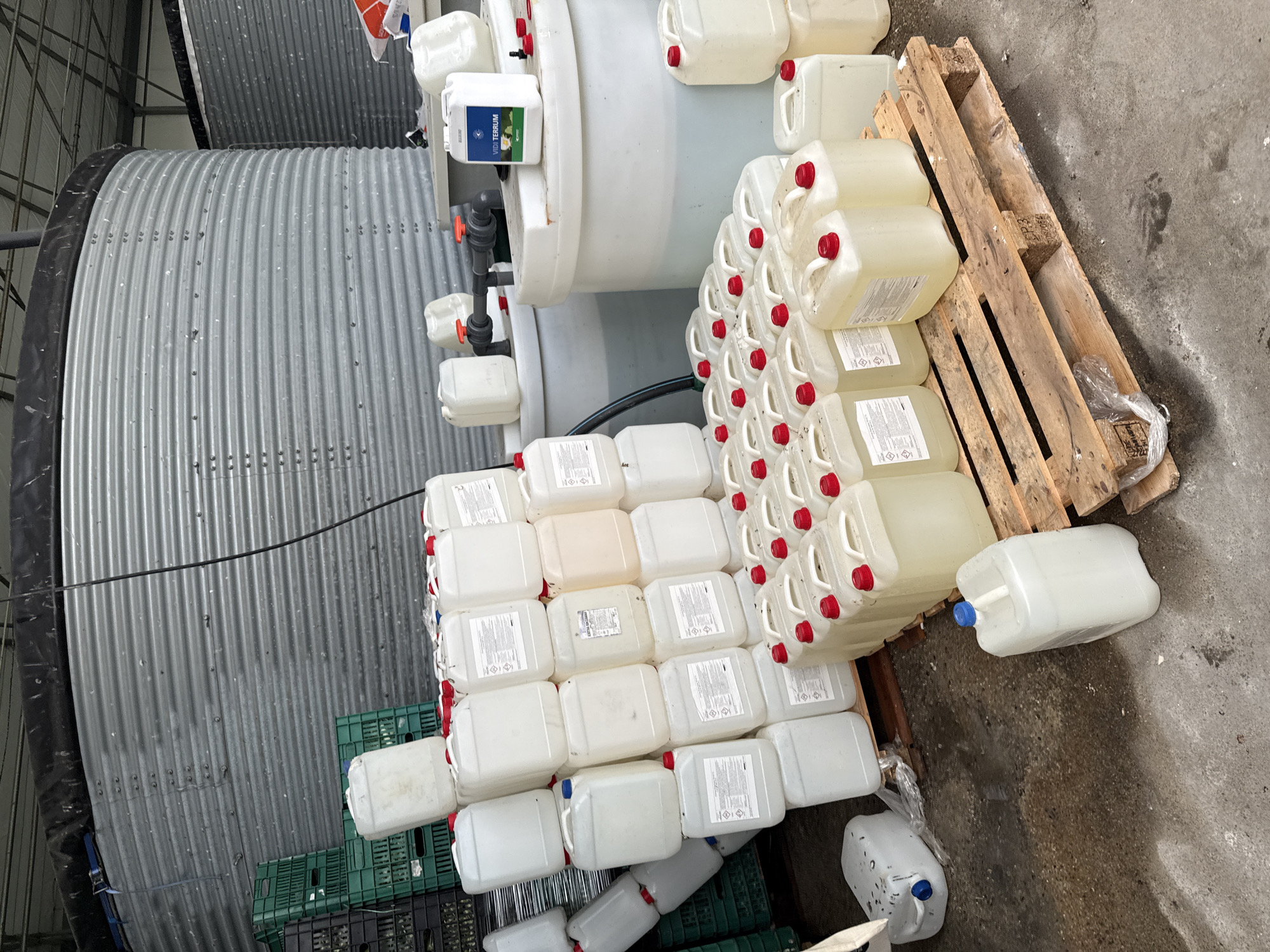 The image shows an indoor scene with various objects. A large, cylindrical, corrugated metal tank with a black top is on the left. Next to it is a white, cylindrical tank with a smaller tank attached. There are white plastic containers with red caps stacked neatly near the tanks. Some containers are also on a wooden pallet. Green plastic crates are visible at the bottom left. The setting appears to be an industrial or agricultural facility, with a concrete floor. The location is Stara Zagora, Bulgaria.