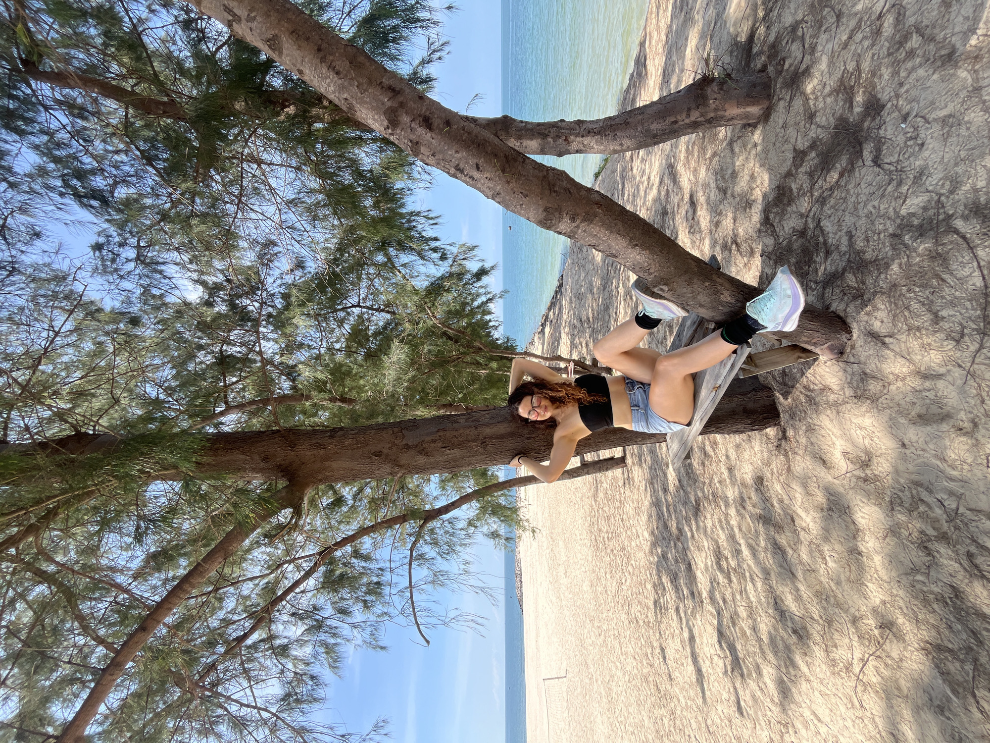 In Phu Thuy Ward, Vietnam, a young woman with curly brown hair and glasses sits on a wooden bench placed between two trees on a sandy beach. She is wearing a black strapless top and denim shorts. The trees are tall with sparse foliage, casting dappled shadows on the sand. The ocean stretches out before her, with a clear blue sky overhead and a calm sea. The beach appears to be mostly empty, with a volleyball net visible in the distance. The woman is looking towards the camera with a slight smile and her hands behind her head.