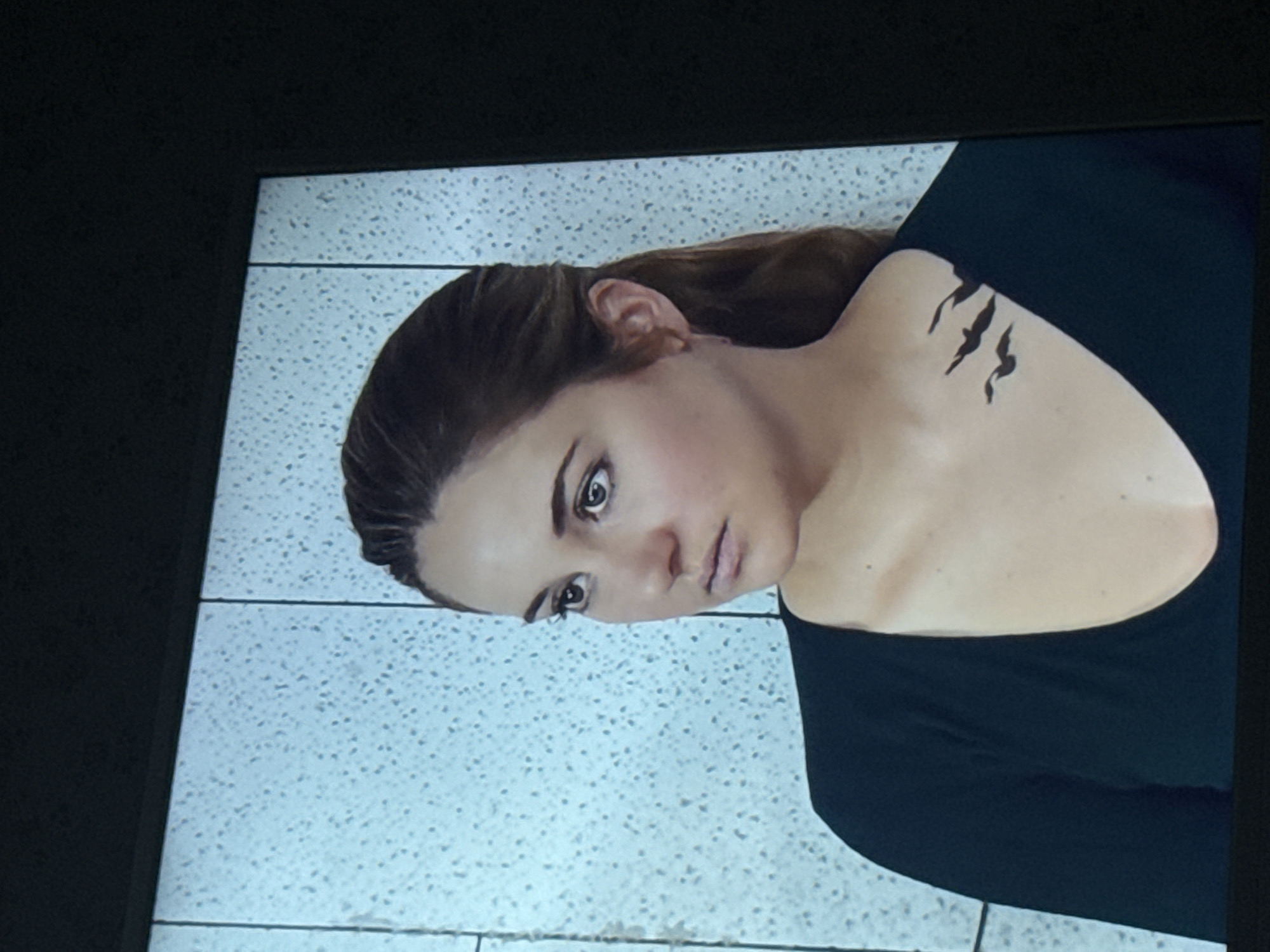 A close-up shot shows a woman with fair skin, brown hair, and dark eyes, wearing a black top. She has a tattoo of what appears to be three claw marks on her upper shoulder. The background appears to be a light-colored, speckled surface with lines suggesting tiling or a similar construction material. There is no information to suggest this image was taken in Mytishchi, Russia.
