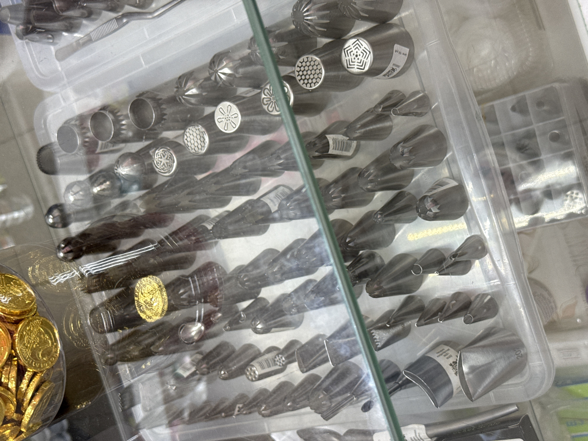 The image displays a close-up, overhead view of a collection of metal piping tips for baking, arranged in clear plastic containers. The piping tips vary in size and design, featuring various shapes such as star tips, round tips, and petal tips, some with intricate patterns. They are primarily silver in color and appear to be made of stainless steel. In the upper left corner of the image, a container filled with gold-foiled chocolate coins is visible. The overall environment suggests a retail setting, possibly a baking supply store or a confectionery shop. The lighting is bright, illuminating the metallic surfaces of the piping tips. The image does not provide any information regarding the city of Lyubertsy, Russia, nor does it depict any people or specific actions.